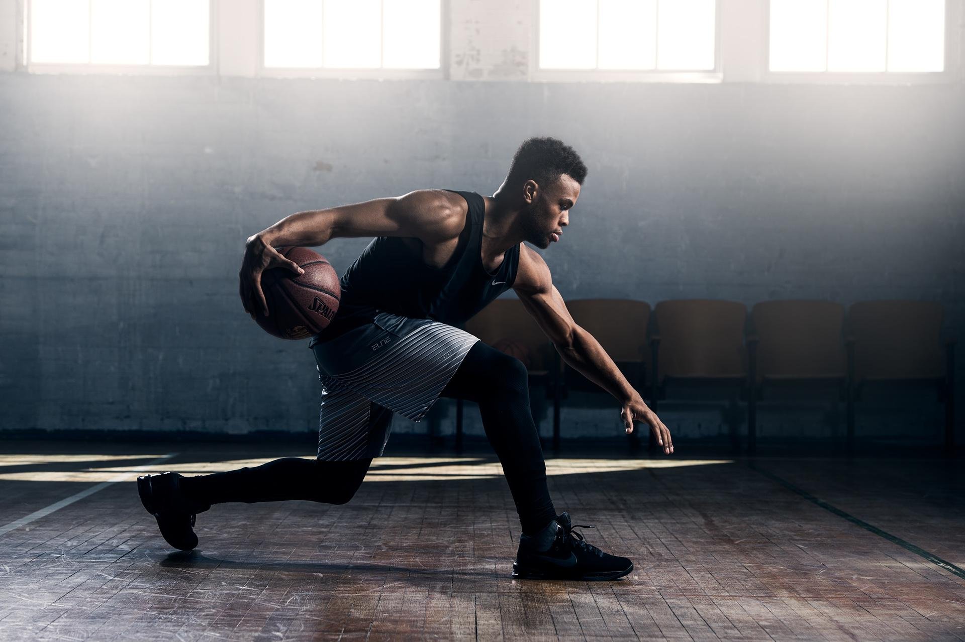 A male athlete in a black sports top and shorts working out with a basketball in an indoor gym, stretching and balancing on one foot, with sunlight coming through windows in the background.