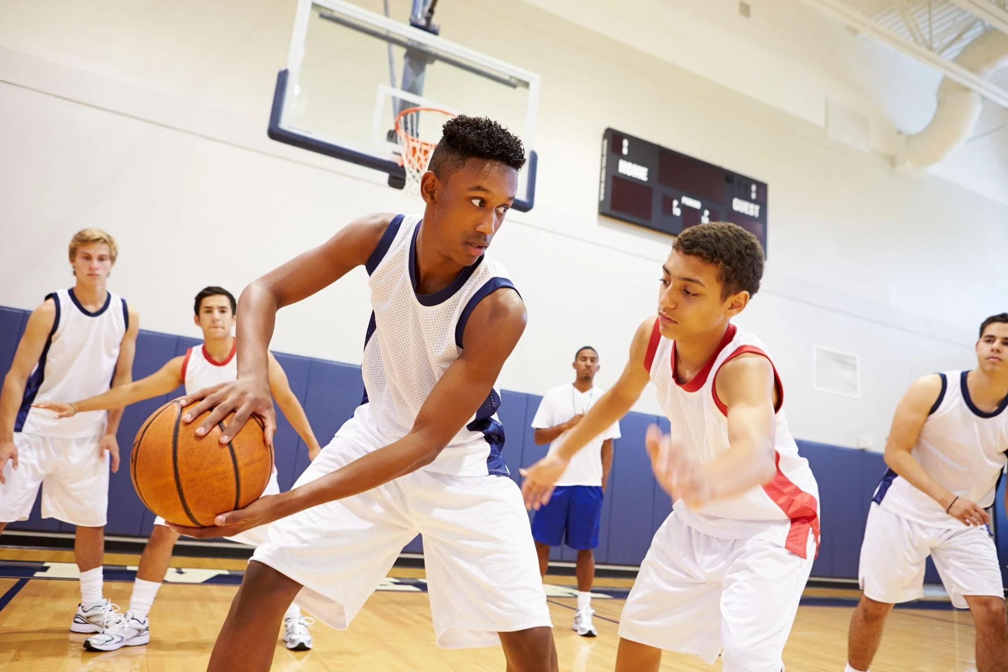 Teen basketball players in an indoor gym playing defense, with one player holding a basketball, while other teens observe and prepare to move.
