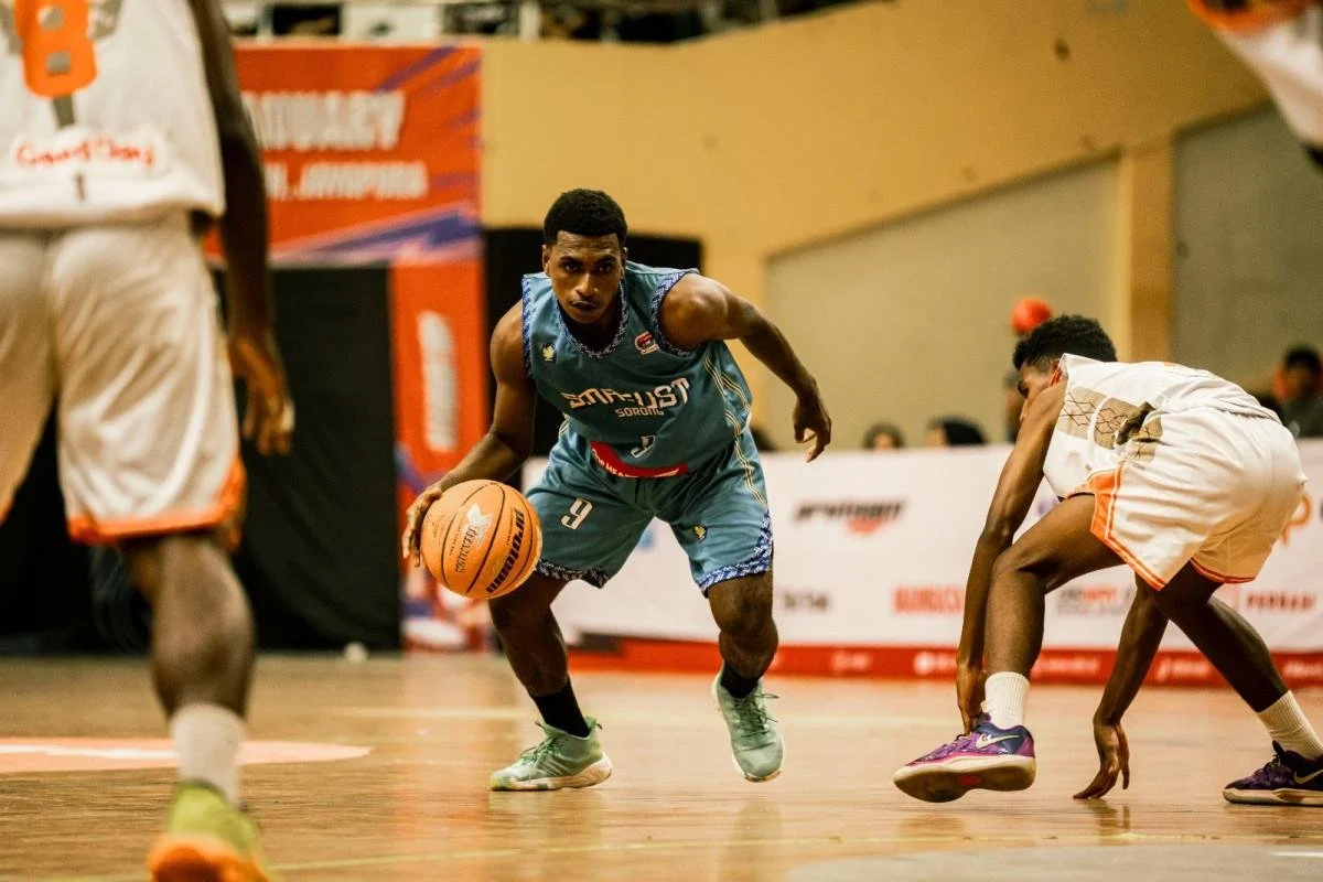 A basketball player in a blue uniform dribbling the ball, while defending against opposing players in white uniforms on a basketball court.