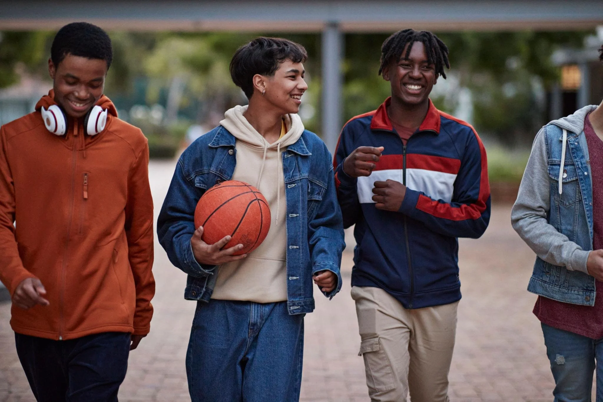 Four young friends walking outdoors, smiling, one holding a basketball, dressed casually.