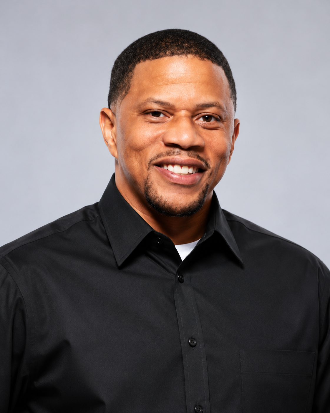 Close-up portrait of a smiling African American man with short hair, beard, and mustache, wearing a black collared shirt against a plain gray background.