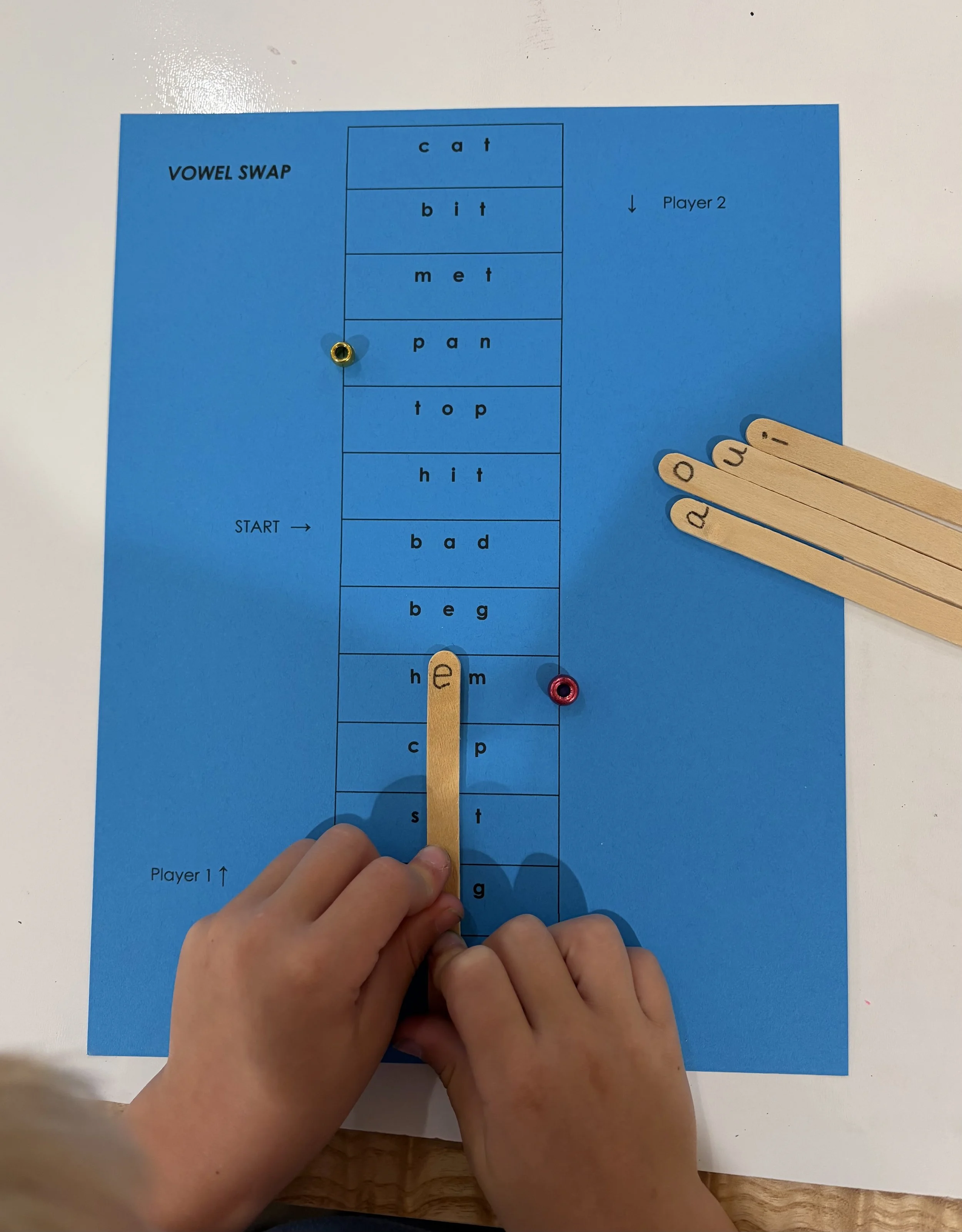 A blue game board with the words 'VOWEL SWAP' at the top left corner, and the words 'Player 1' and 'Player 2' at the bottom left and right corners, respectively. The board displays columns with various words featuring vowels, such as 'cat,' 'bit,' 'met,' 'pan,' 'top,' 'hit,' 'bad,' and 'beg,' with some corresponding wooden game pieces placed on certain words and a hand holding a wooden piece near the middle of the board.
