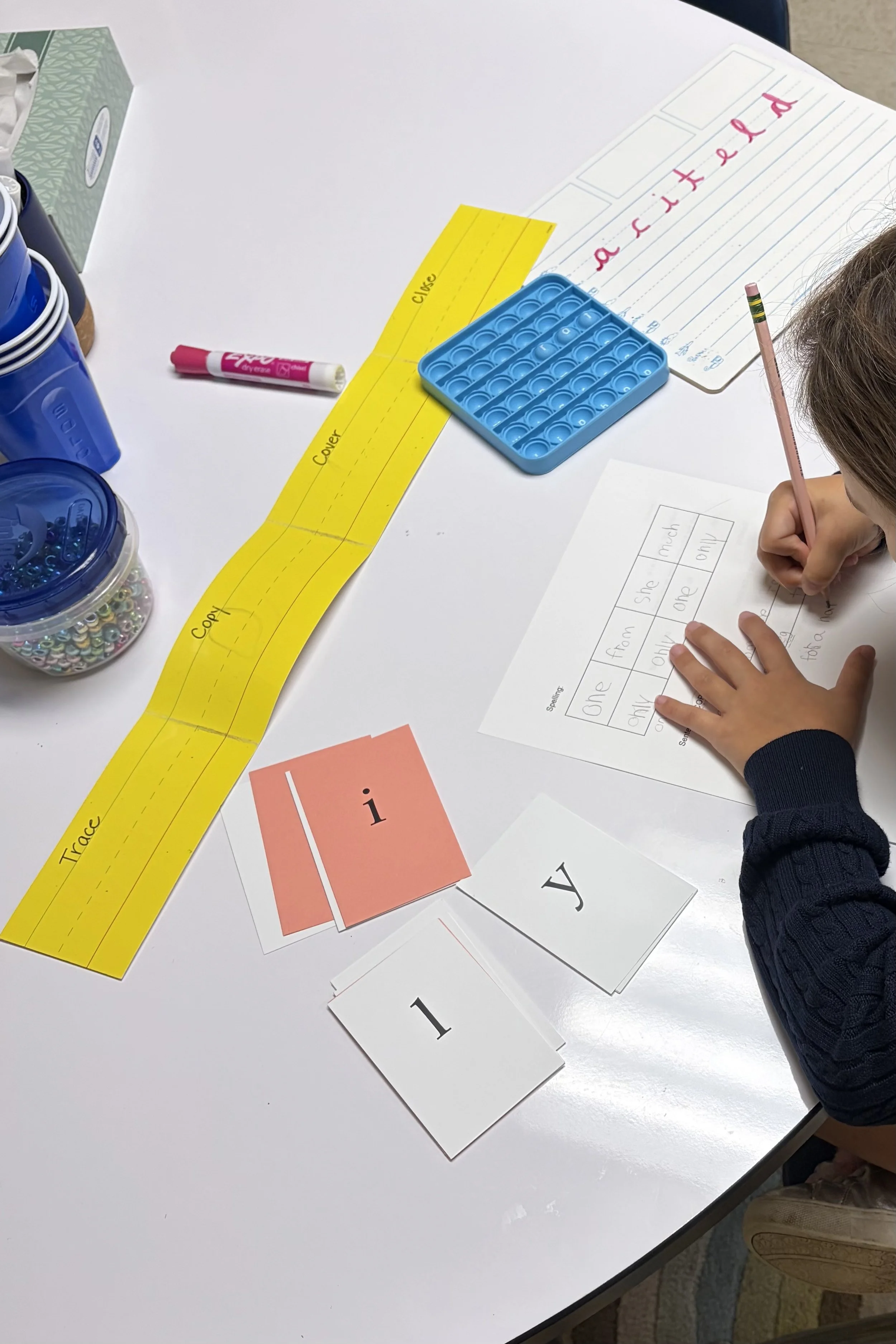 A child's hand holding a pink pencil writing on a lined classroom worksheet with words and pictures. Various educational materials, including color-coded word cards, a yellow word strip, and small containers of alphabet letter decals, are spread out on a white table.