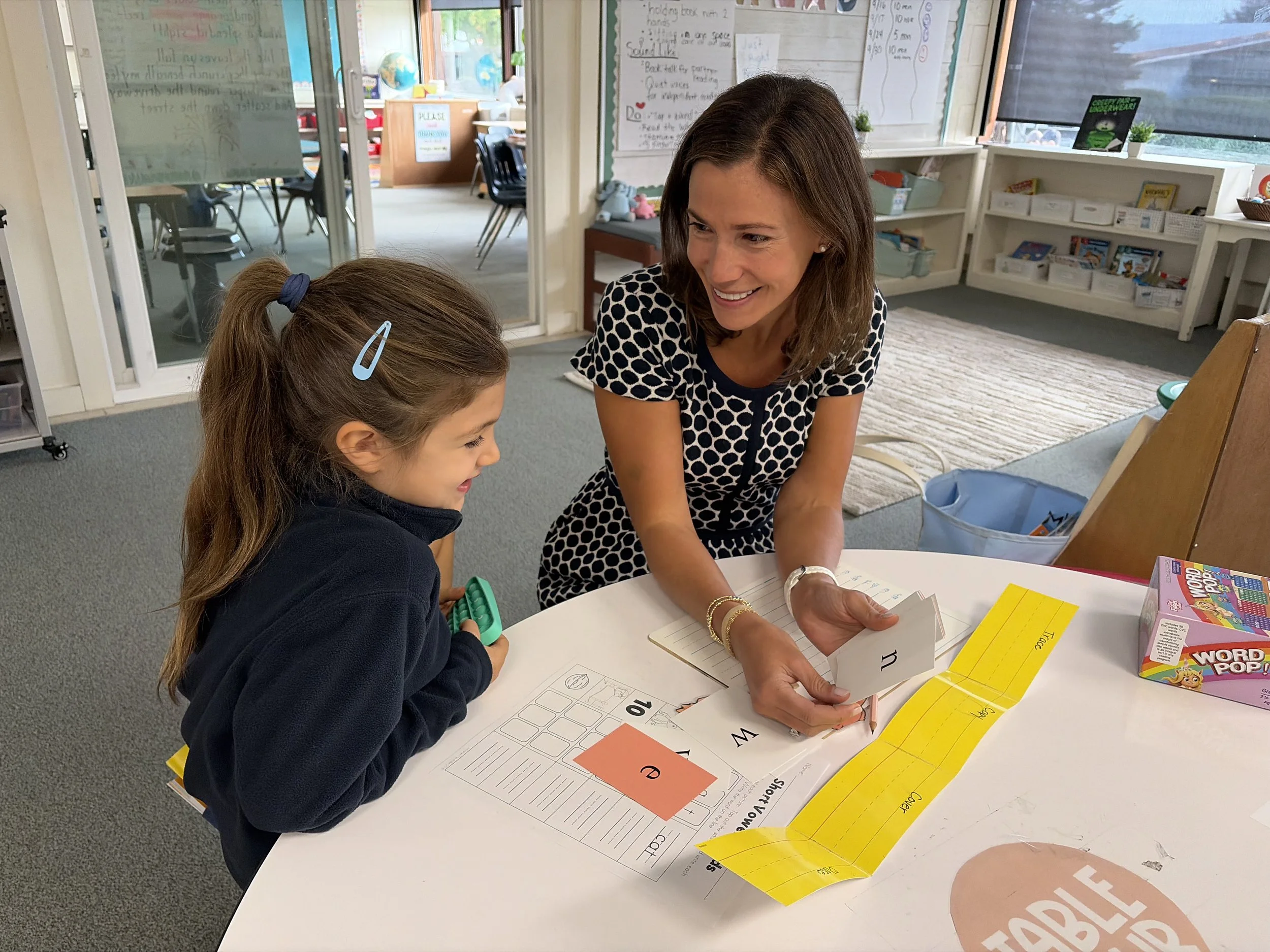 A woman smiling at a young girl during a classroom activity. The girl has long brown hair in a ponytail and is holding a green object. They are sitting at a white table with educational materials and colorful papers. The background shows bookshelves, posters, and a glass door leading to another room.