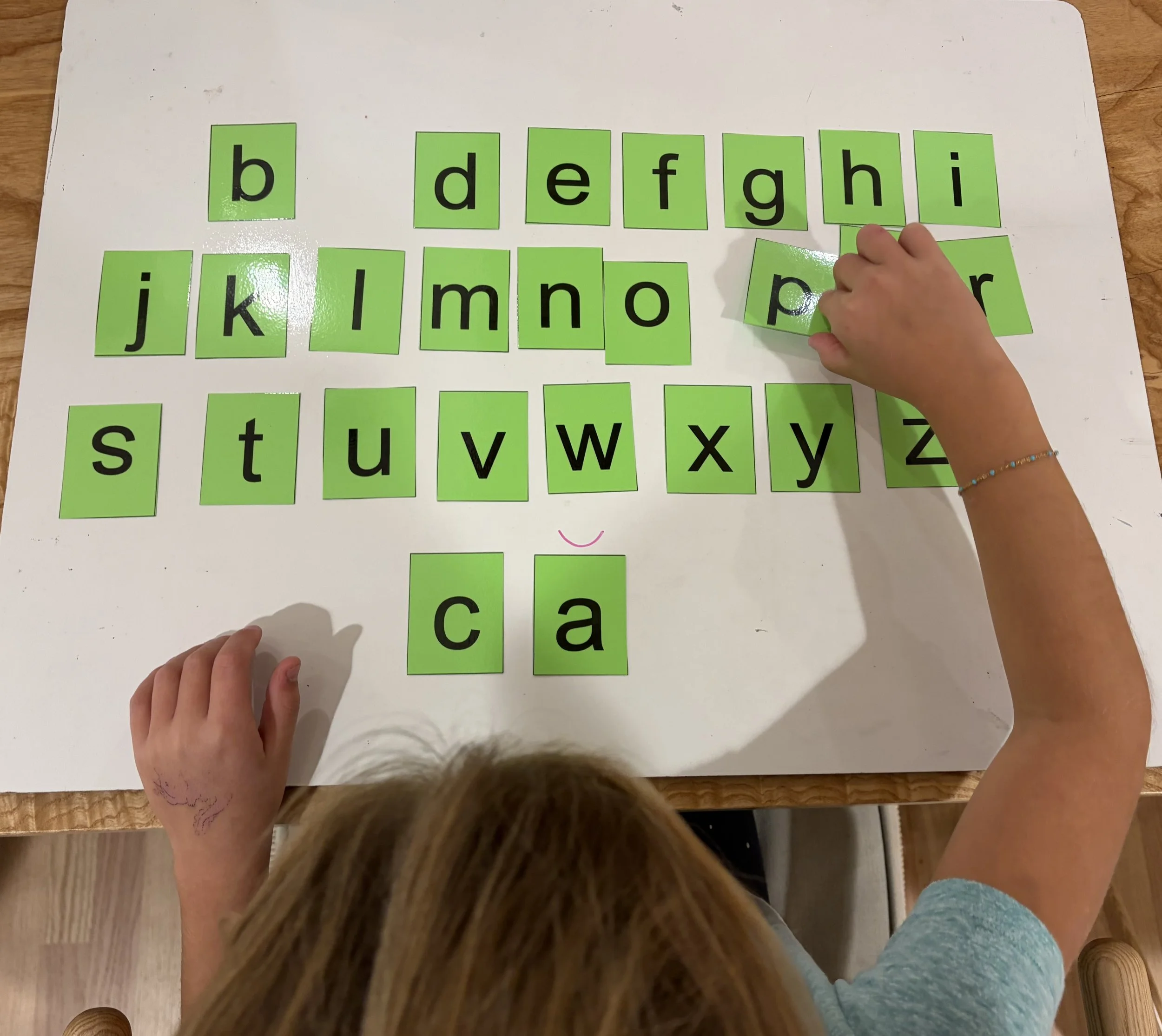 Children's hands arranging green flashcards with lowercase letters on a wooden table.