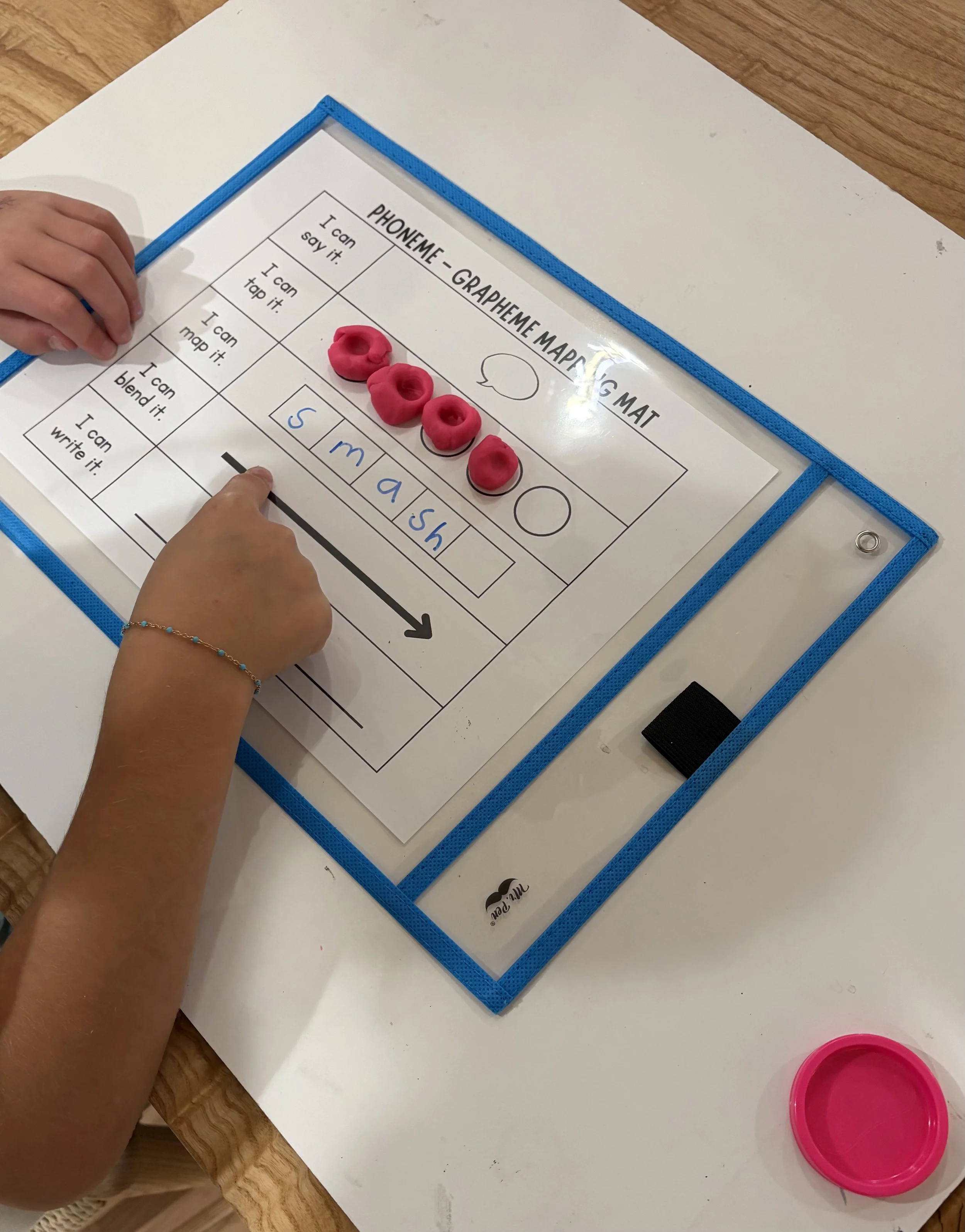Children's hands working on a phoneme grapheme mapping mat with pink playdough letters and blue written words, on a white table.