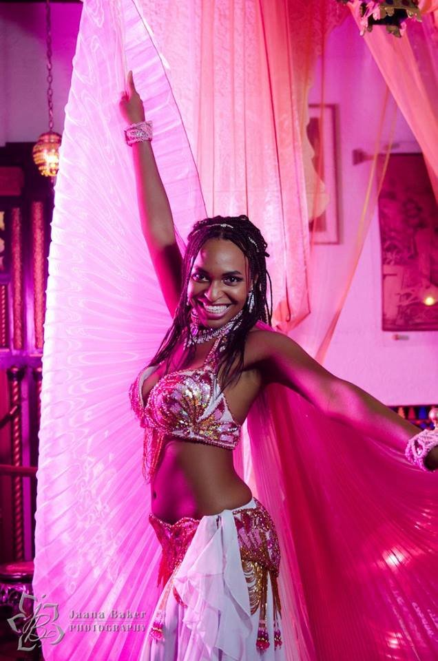 A woman dressed in colorful, shiny belly dance costume with a bejeweled top and skirt, holding a large pink fan, smiling in a decorated venue with pink and purple lighting.