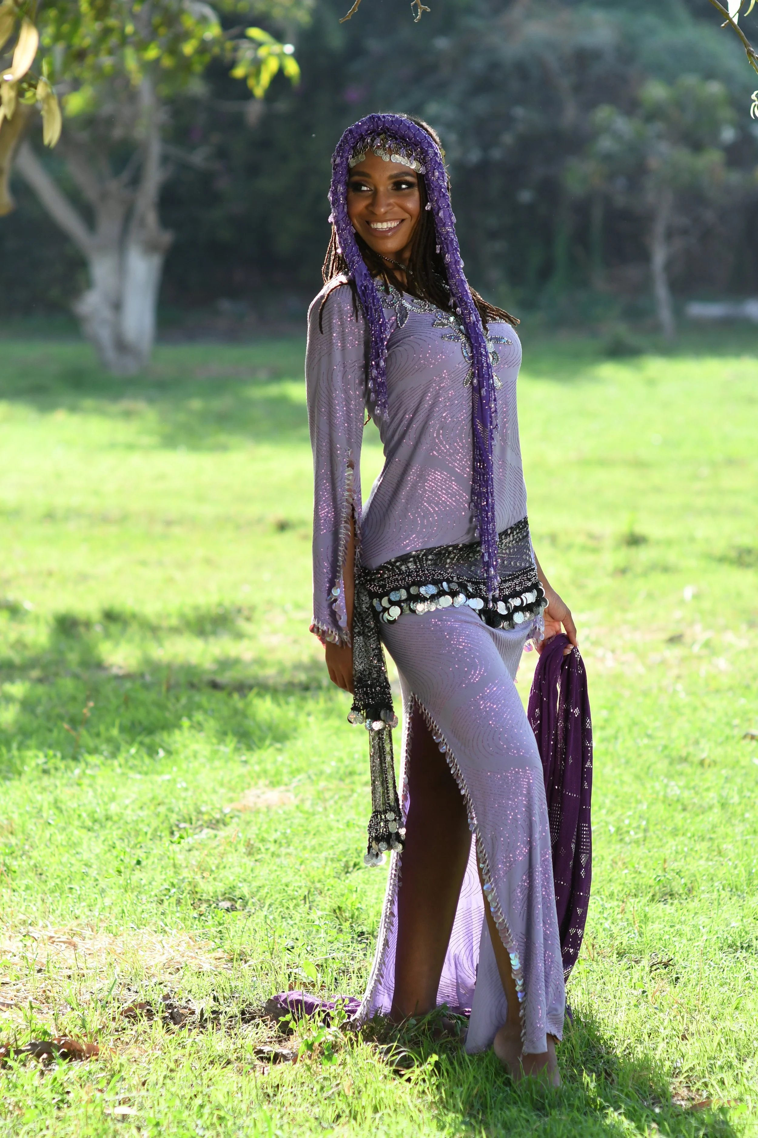 A woman dressed in a shiny purple costume with silver embellishments and a matching purple headpiece, standing outdoors on a grassy area with trees in the background, smiling at the camera.
