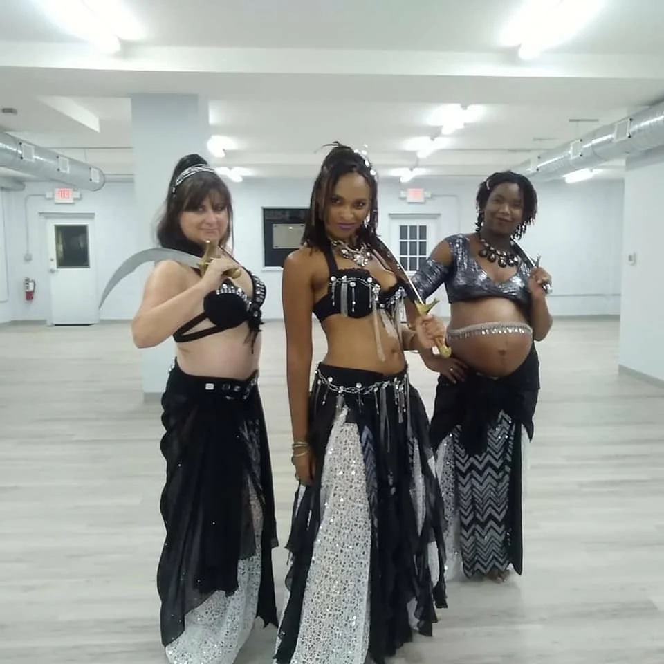 Three women in black and silver costume standing in an empty room, each holding a curved sword and dressed in tribal or fantasy-inspired attire.