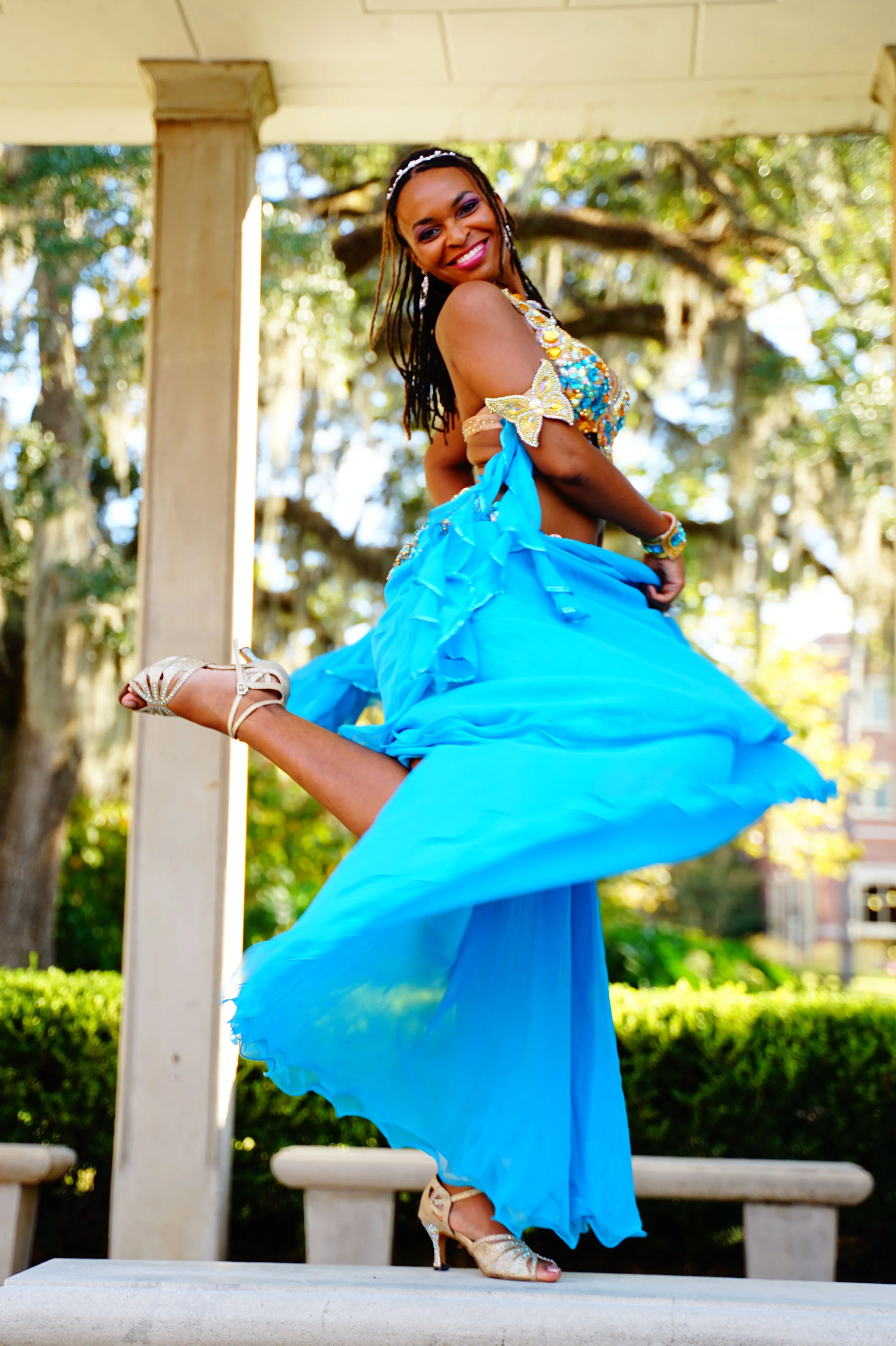 A woman in a bright blue dress with embellishments on the top poses with one leg raised and her hands on her waist outdoors under a porch, surrounded by trees and greenery.