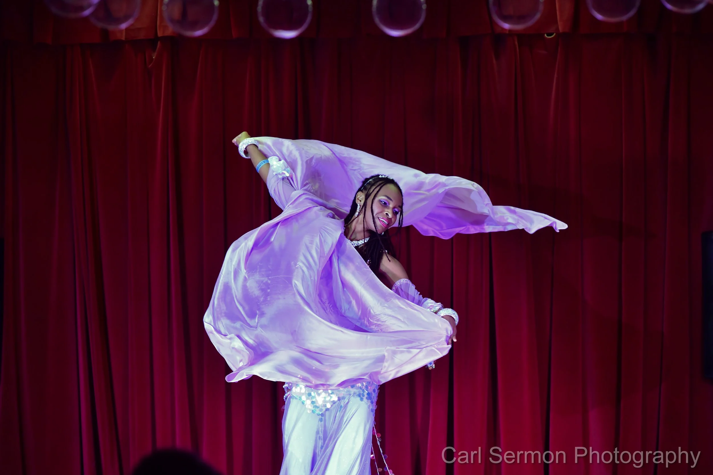A dancer performing on stage in front of a red curtain, wearing a shiny, iridescent outfit and spinning with a flowing purple fabric.