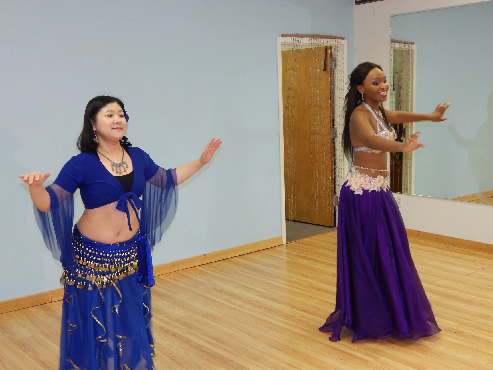 Two women performing a dance in a room with a wooden floor, one wearing a royal blue costume with gold embellishments and the other wearing a purple outfit with floral decorations, both with arms extended.