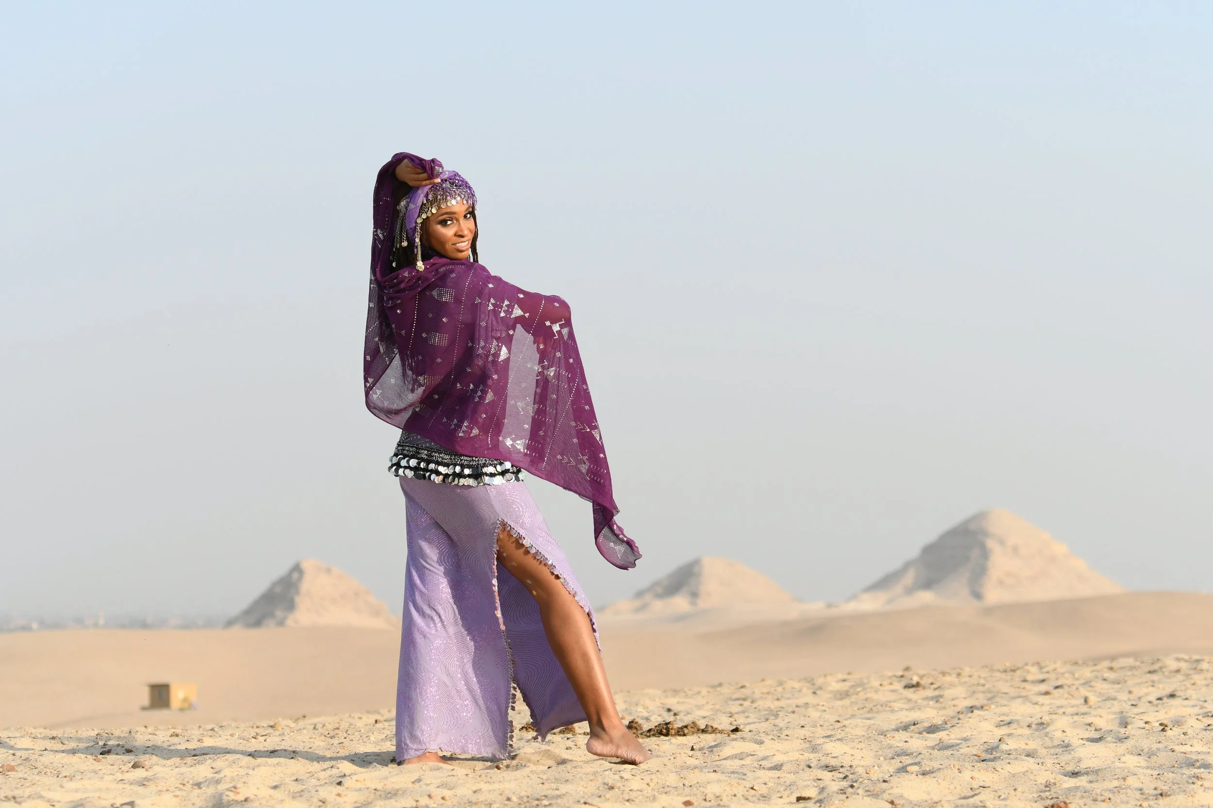 A woman in purple traditional Middle Eastern attire standing barefoot on desert sand with pyramids in the background.