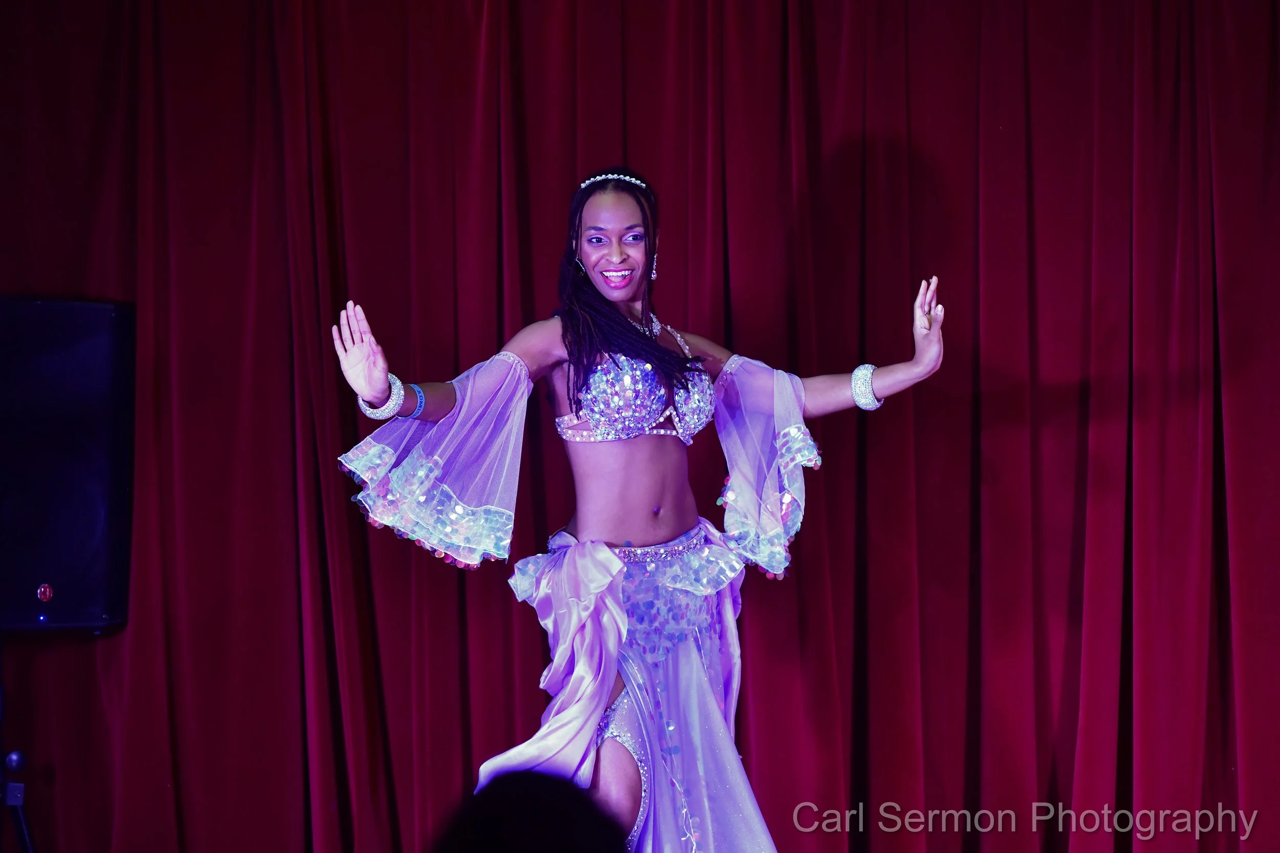 A woman in a sparkling purple and pink belly dancing costume performing on stage with red curtains in the background.