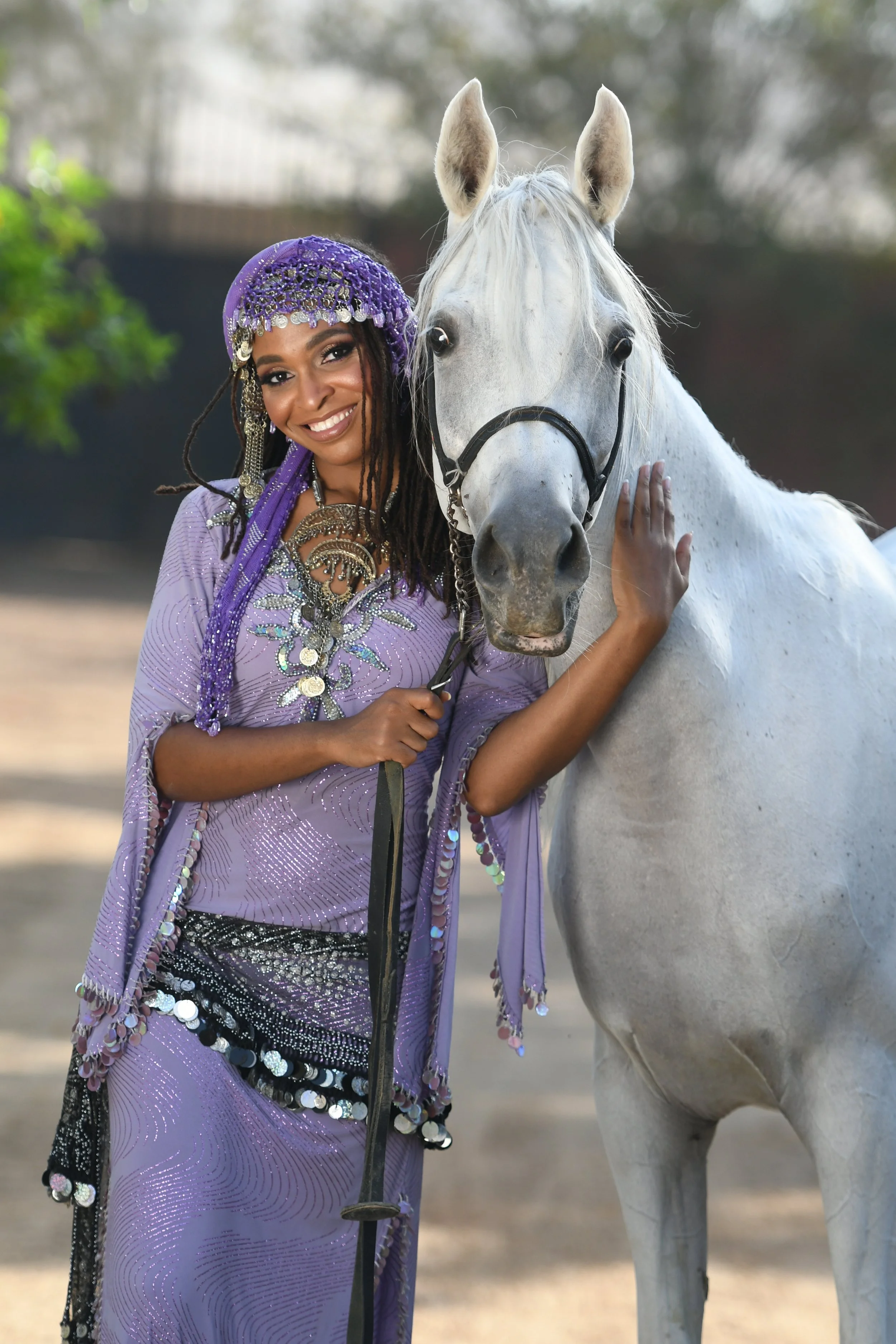A woman dressed in a purple, sequin-adorned outfit with beads and jewelry is standing next to a white horse, touching its face gently, outdoors with blurred trees in the background.