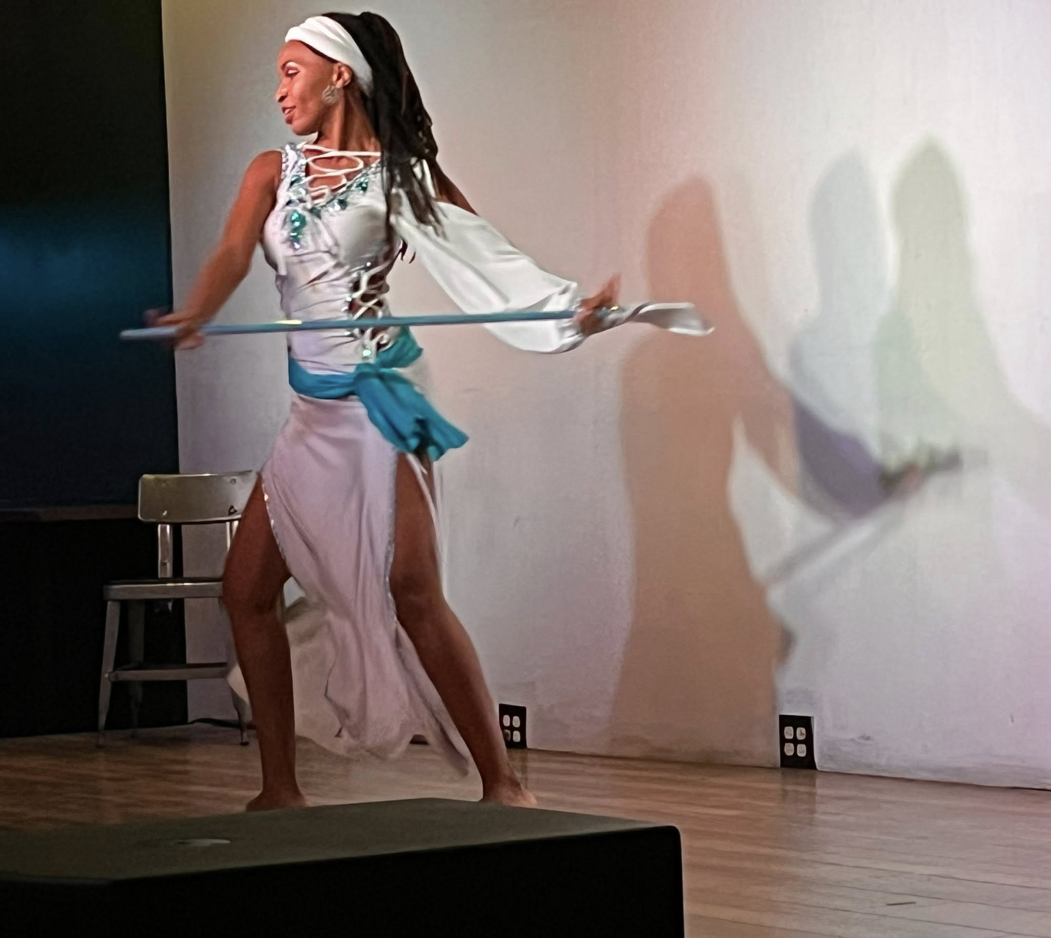 A woman with long dark hair, wearing a white headband and a white dress with cut-out details, is standing barefoot on a wooden floor near a white wall, performing or practicing a dance or pose. Her shadow is cast on the wall.