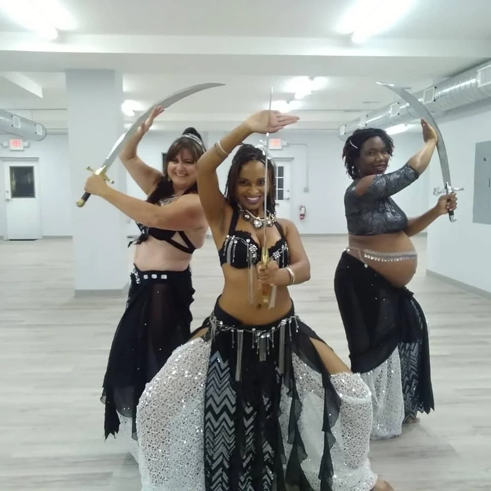 Three women in belly dance costumes holding swords, posing in a dance studio.