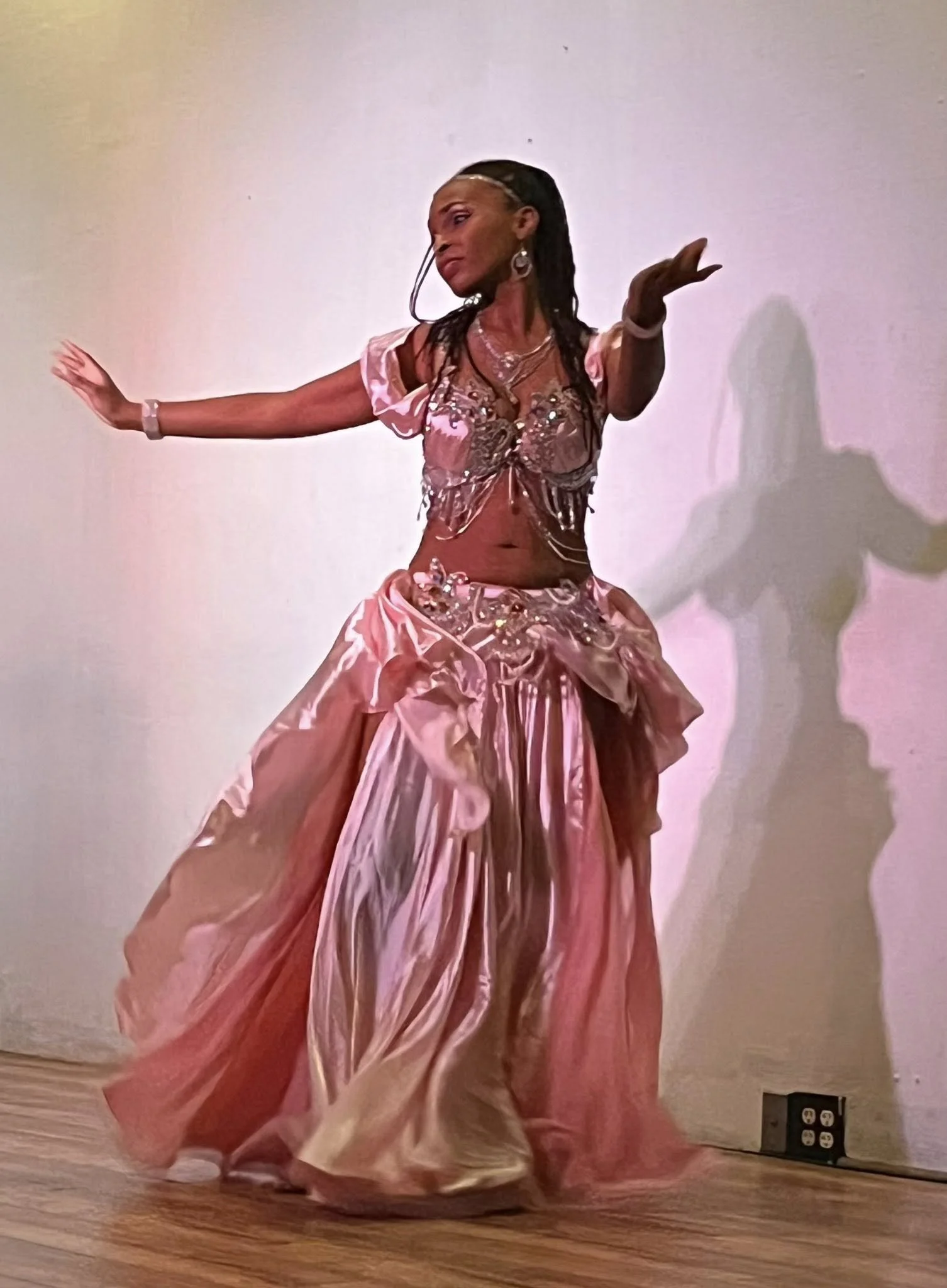 A woman in a pink, flowing, elaborate dress performing a dance on a wooden floor, with a white wall background.