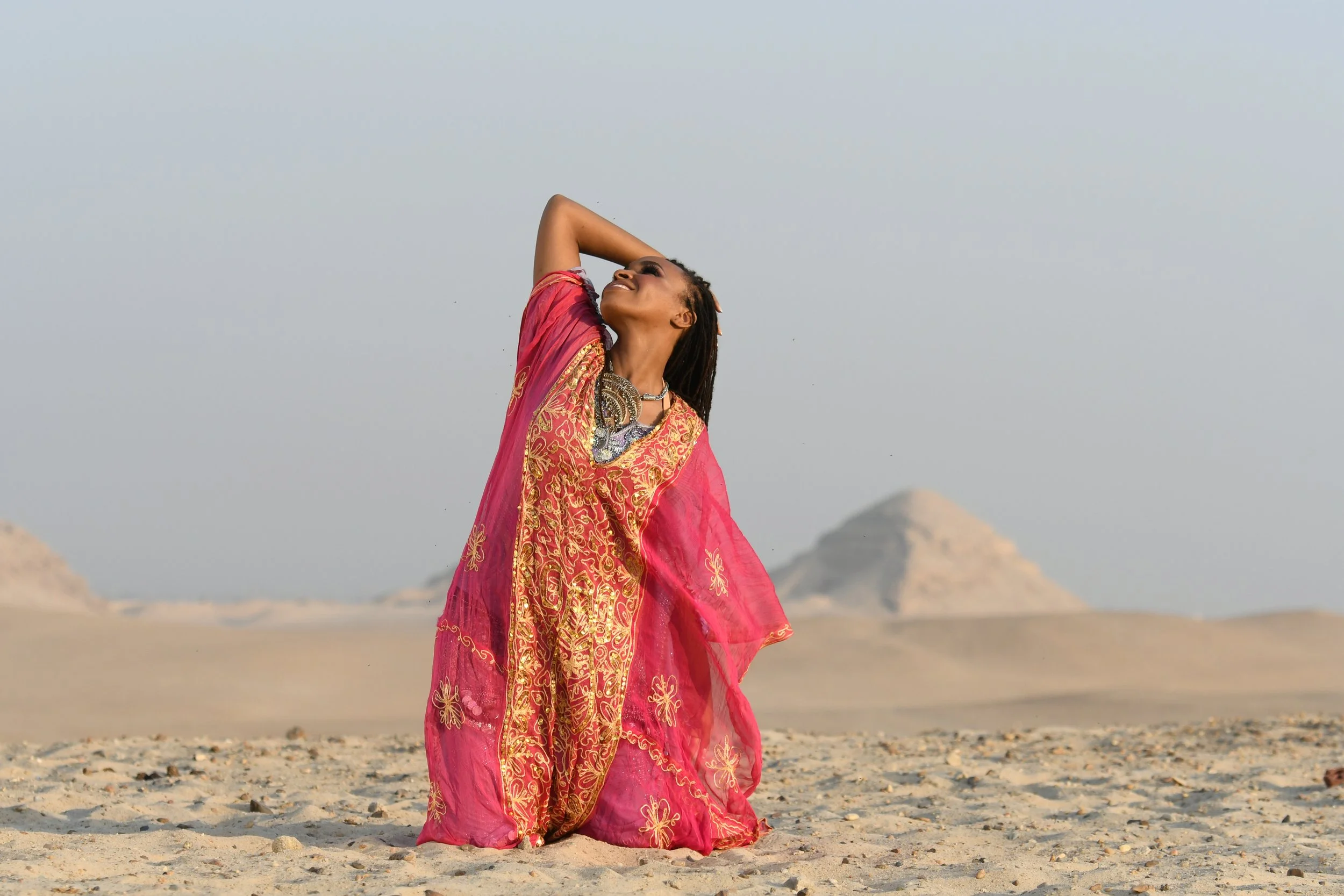 A woman dressed in a vibrant pink traditional dress with gold embroidery, standing on a sandy desert landscape with distant mountains and a cloudy sky, posing with one arm raised above her head and smiling.