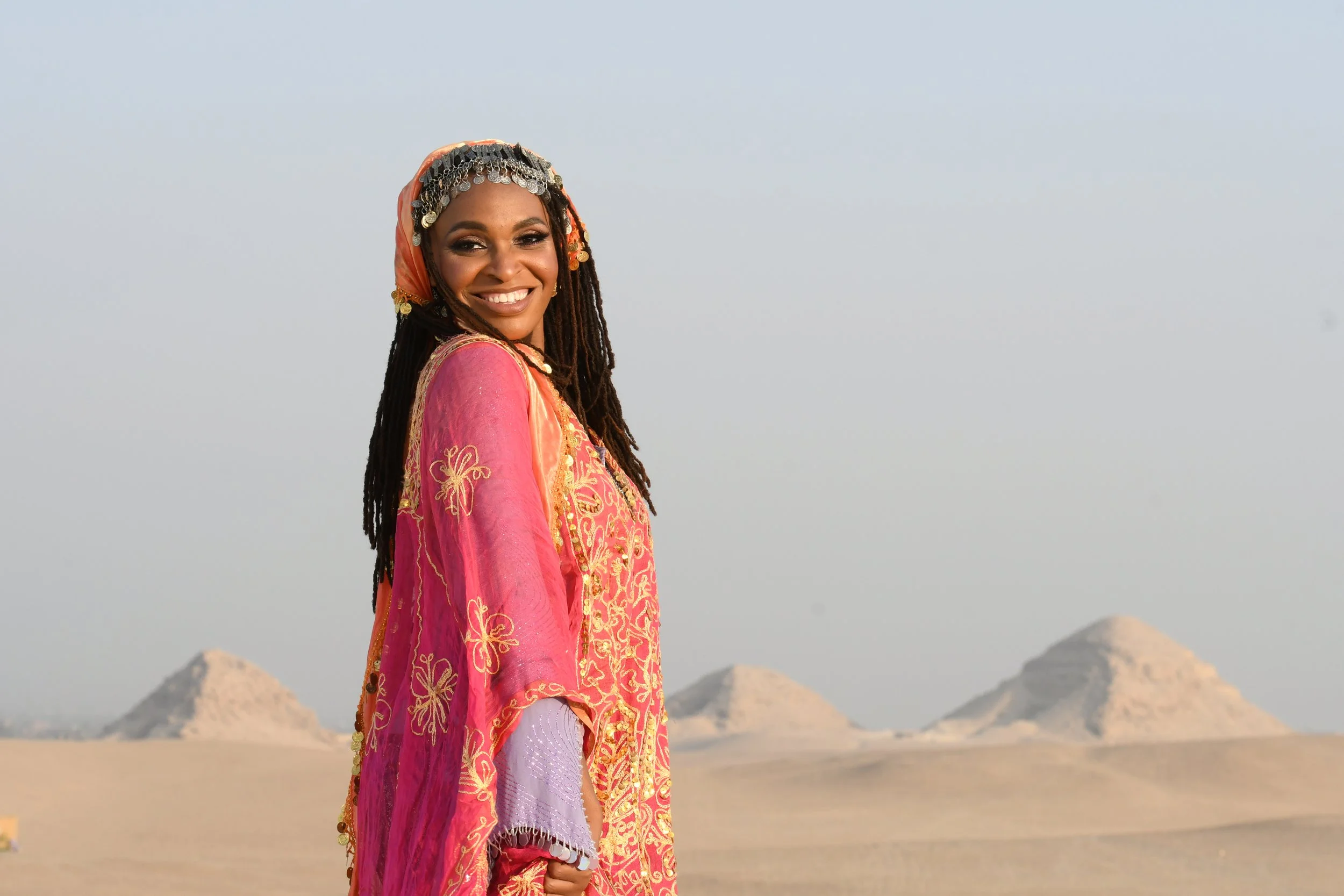 A woman in colorful traditional attire smiling in a desert landscape with pyramids in the background.