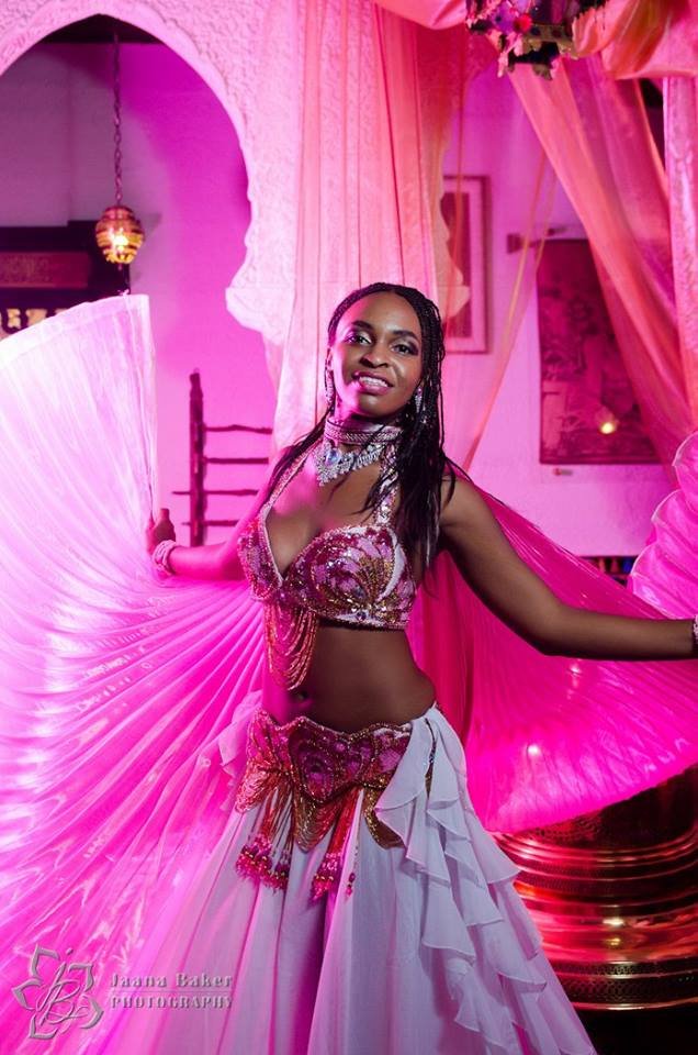A woman in a pink costume with sequins and jewelry, holding a large fan in a brightly lit room with pink lighting, curtains, and decorative elements.