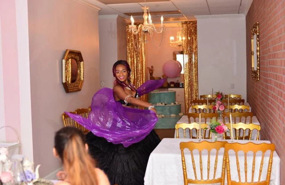 A woman dancing in a party room with gold chairs, floral centerpieces, a chandelier, and gold curtains.