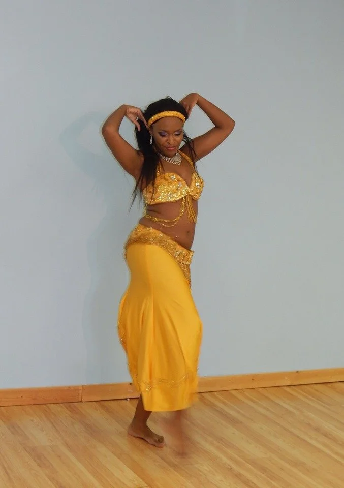 Woman in a yellow belly dance costume with jewelry, standing barefoot on a hardwood floor against a plain wall, striking a dance pose with her hands raised behind her head.