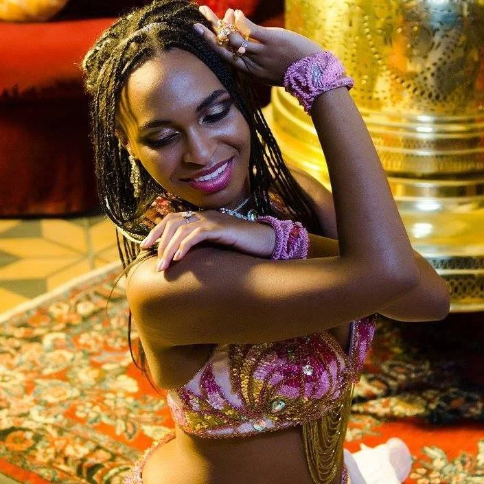 A woman with long braided hair, smiling with her eyes closed, in a colorful outfit, wearing jewelry, and standing indoors near a large decorative plant.