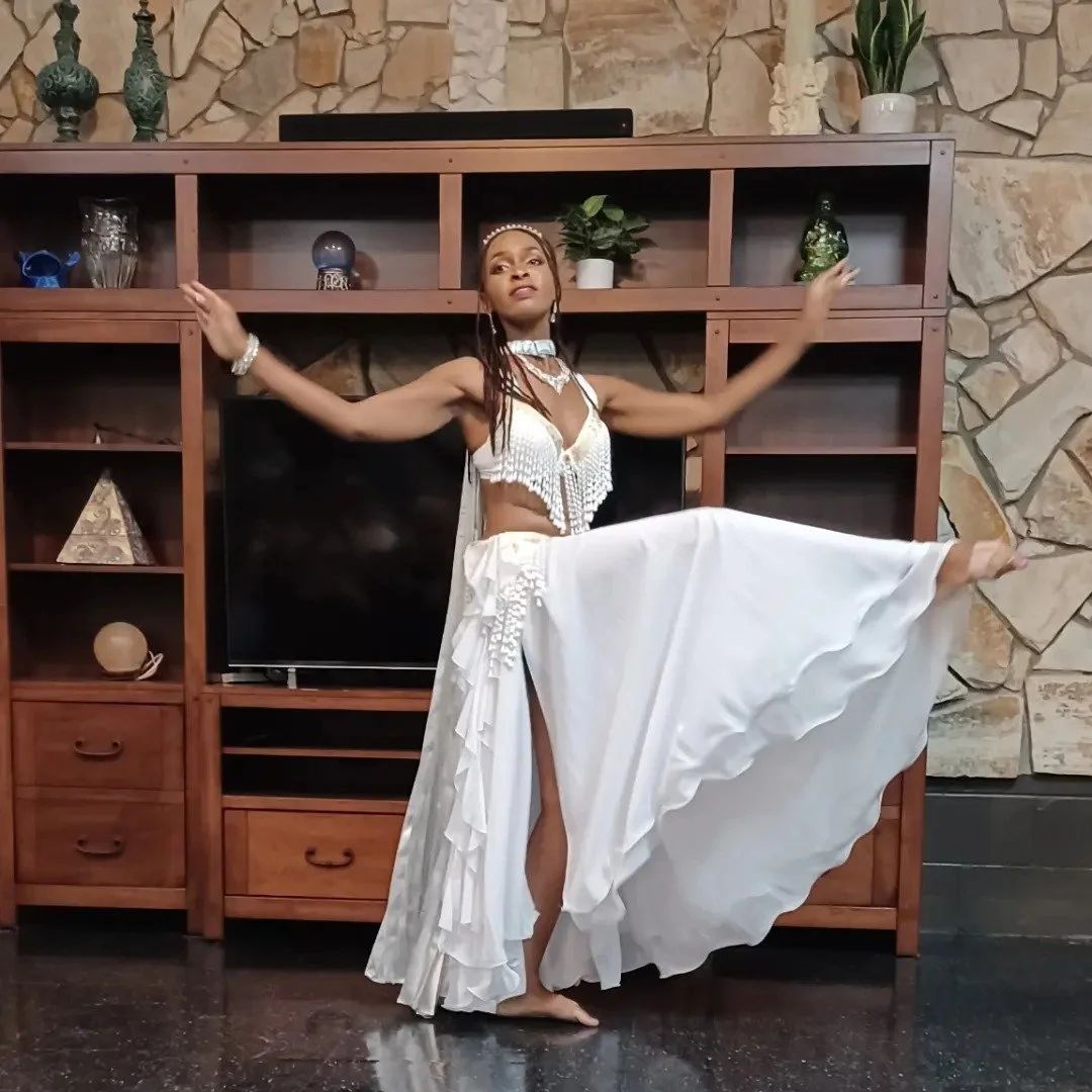 A woman in a white dance costume with fringe and ruffles performs a dance pose in front of a wooden entertainment center with decorative items and a stone wall background.