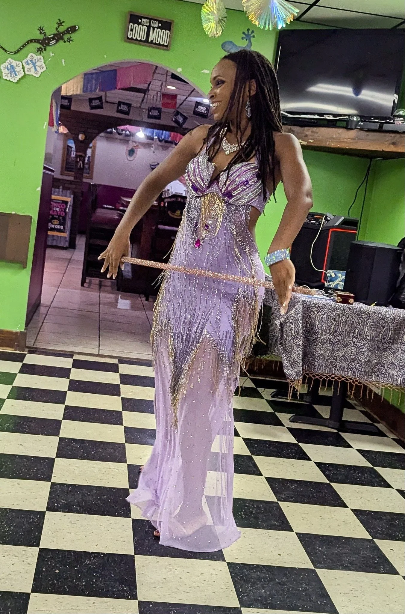 A woman in a purple, beaded gown with gold accents and fringe, smiling and standing on a black-and-white checkered floor inside a restaurant or event space with green walls and holiday decorations.