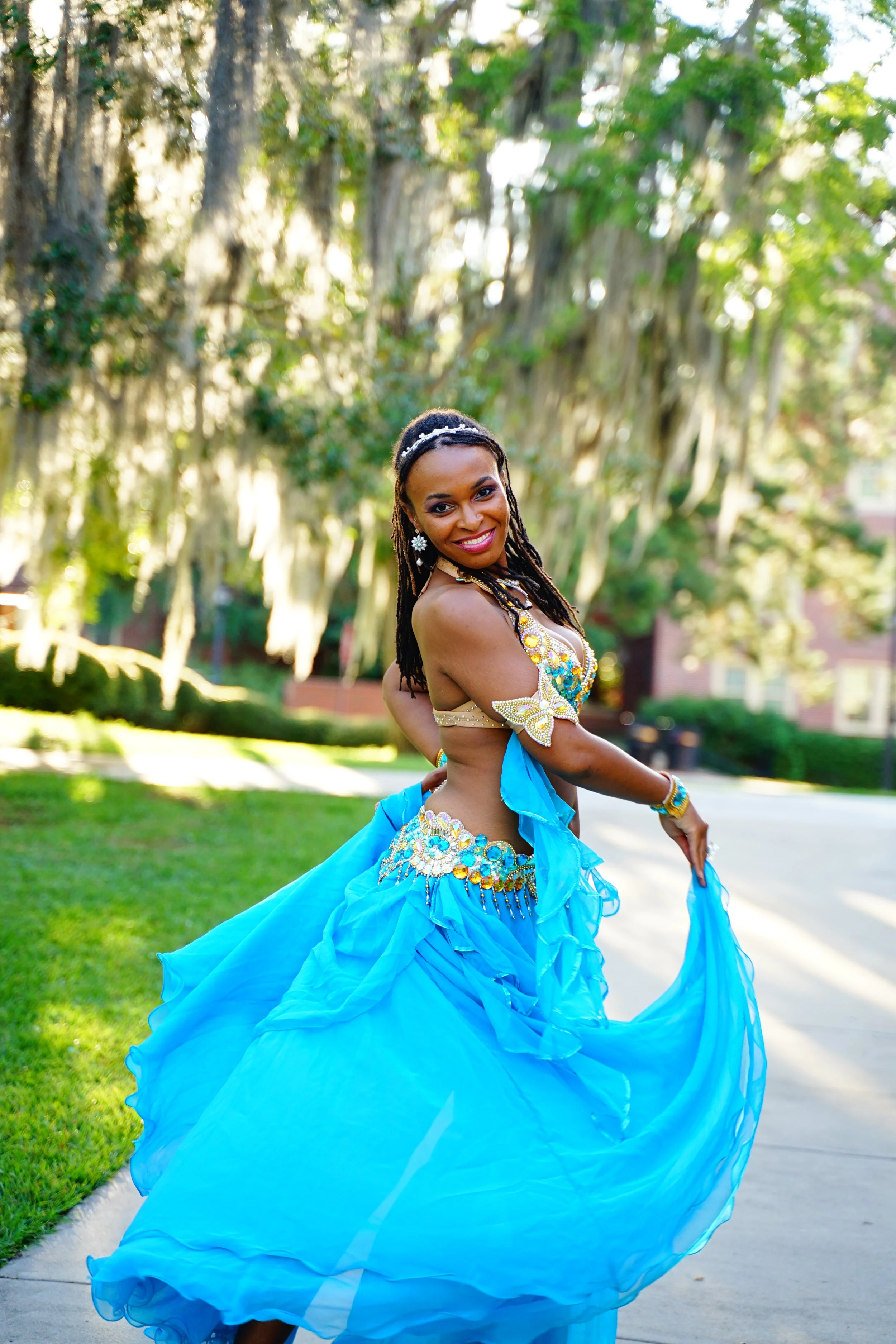 A woman in a vibrant blue belly dance costume dancing outdoors in a park with trees and sunlight in the background.