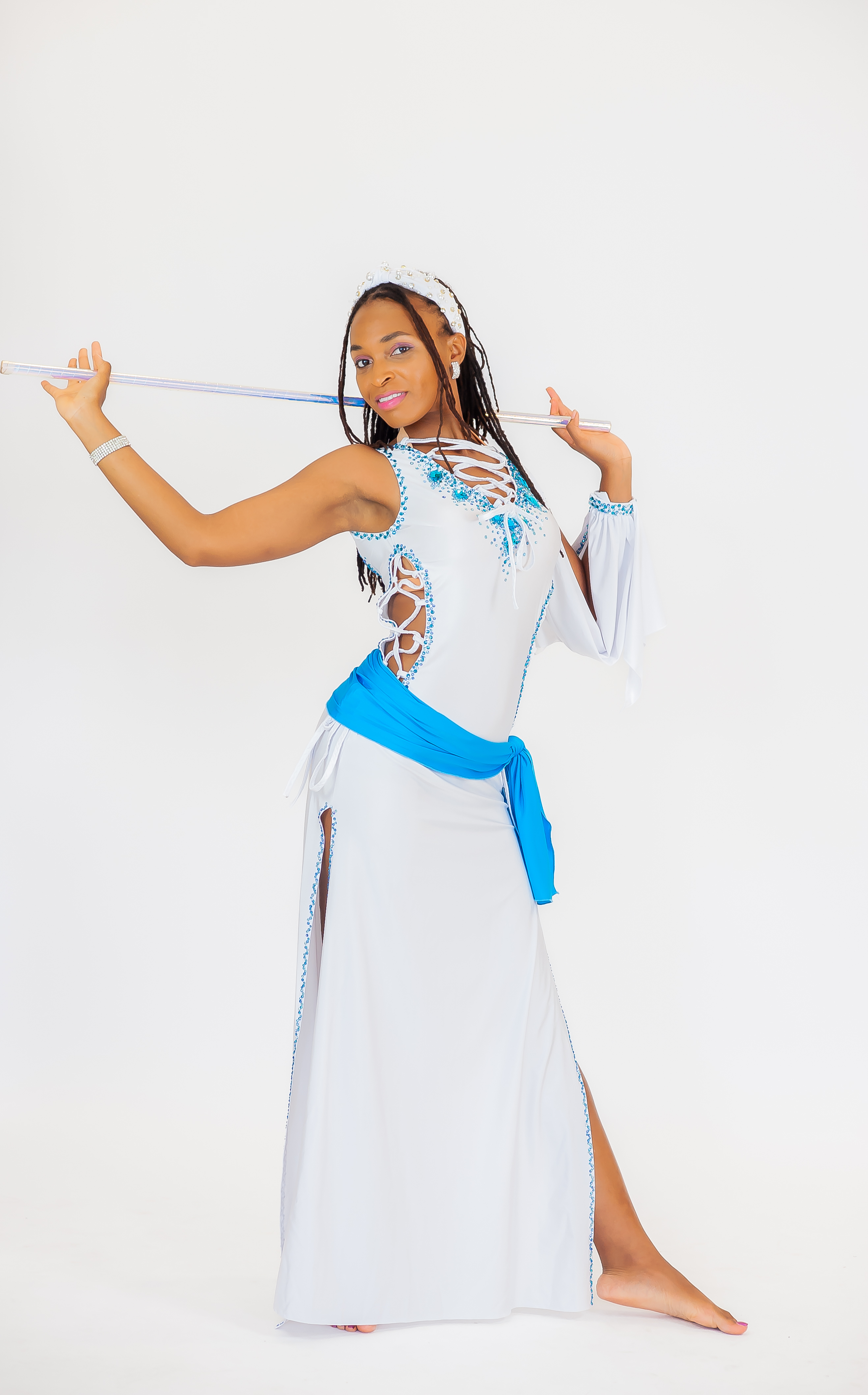 A woman in a white dress with blue accents, holding a long stick over her shoulder, posing against a plain white background.