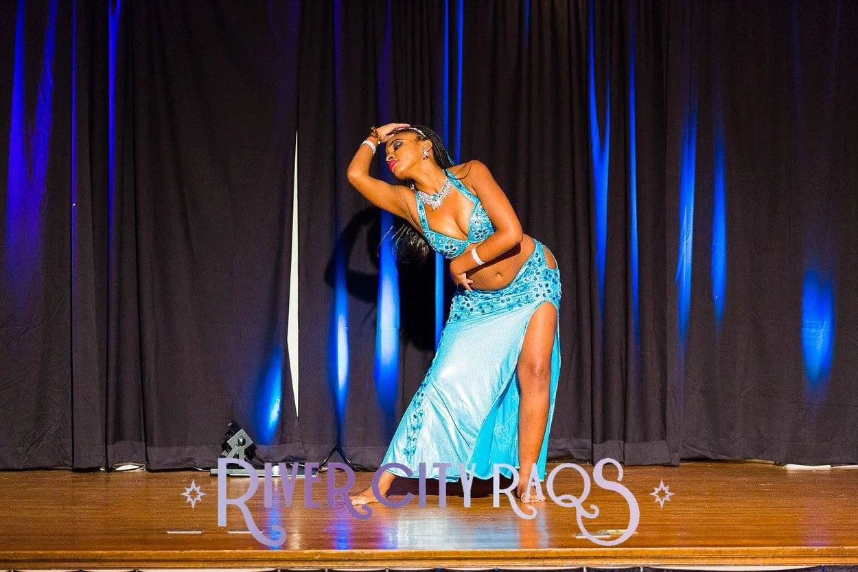 A woman performing a dance on stage in a blue costume with cutouts and high slit, holding her head with one hand and striking a pose, with a dark curtain and blue lights in the background, and a sign reading "River City Rags" on the stage floor.