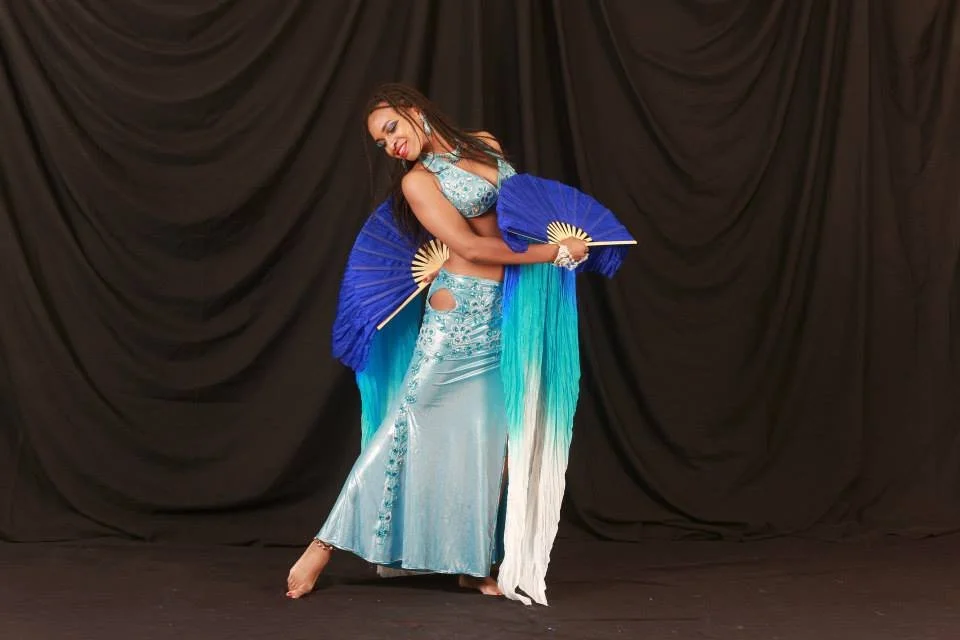 A woman in a blue and silver belly dance costume holding two blue fans stands on a stage with a black curtain backdrop.