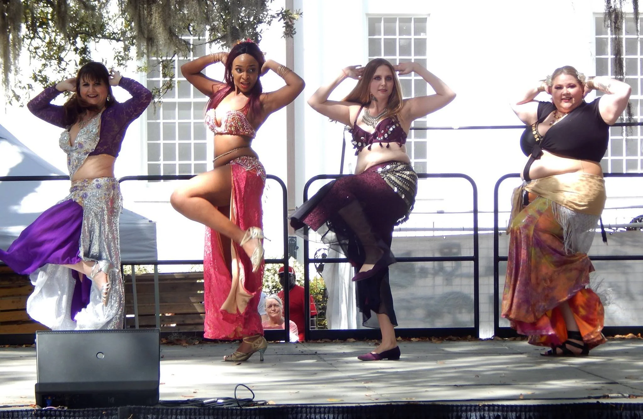 Four women in colorful clothing performing a dance on a stage outdoors, with trees and a white building in the background.