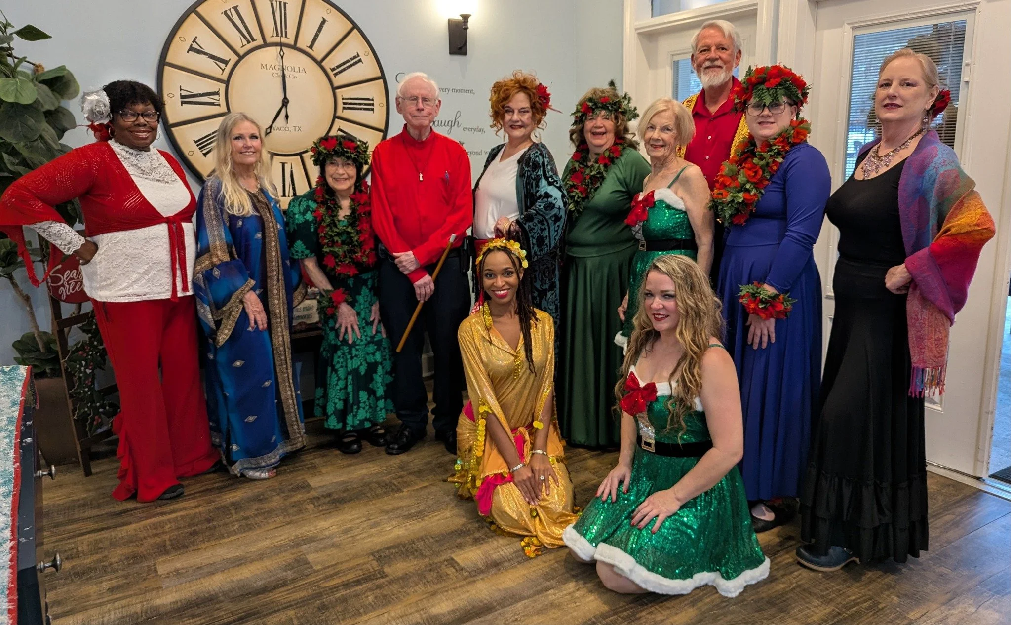 A group of twelve people, dressed in colorful, festive clothing with some wearing flower crowns, standing in a room with a large clock on the wall, celebrating a holiday or special occasion.