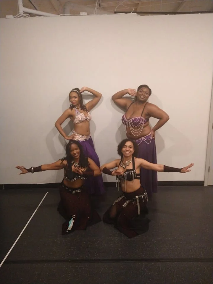 Four women in dance costumes posing in front of a plain white wall. Two women are standing at the back, and two women are kneeling in front, all striking poses.