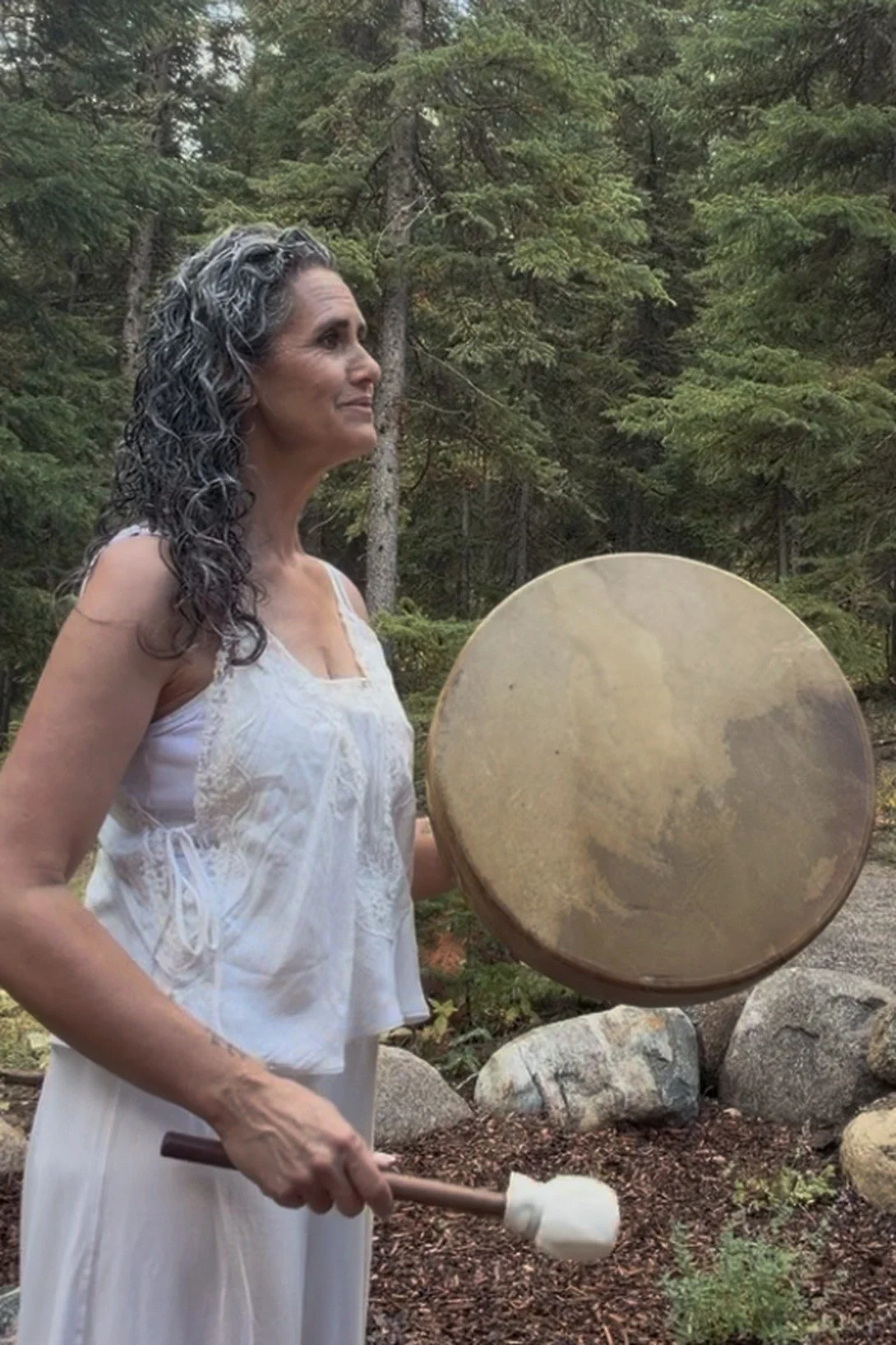 Alana playing a frame drum in a Montana pine forest during an outdoor sound healing session