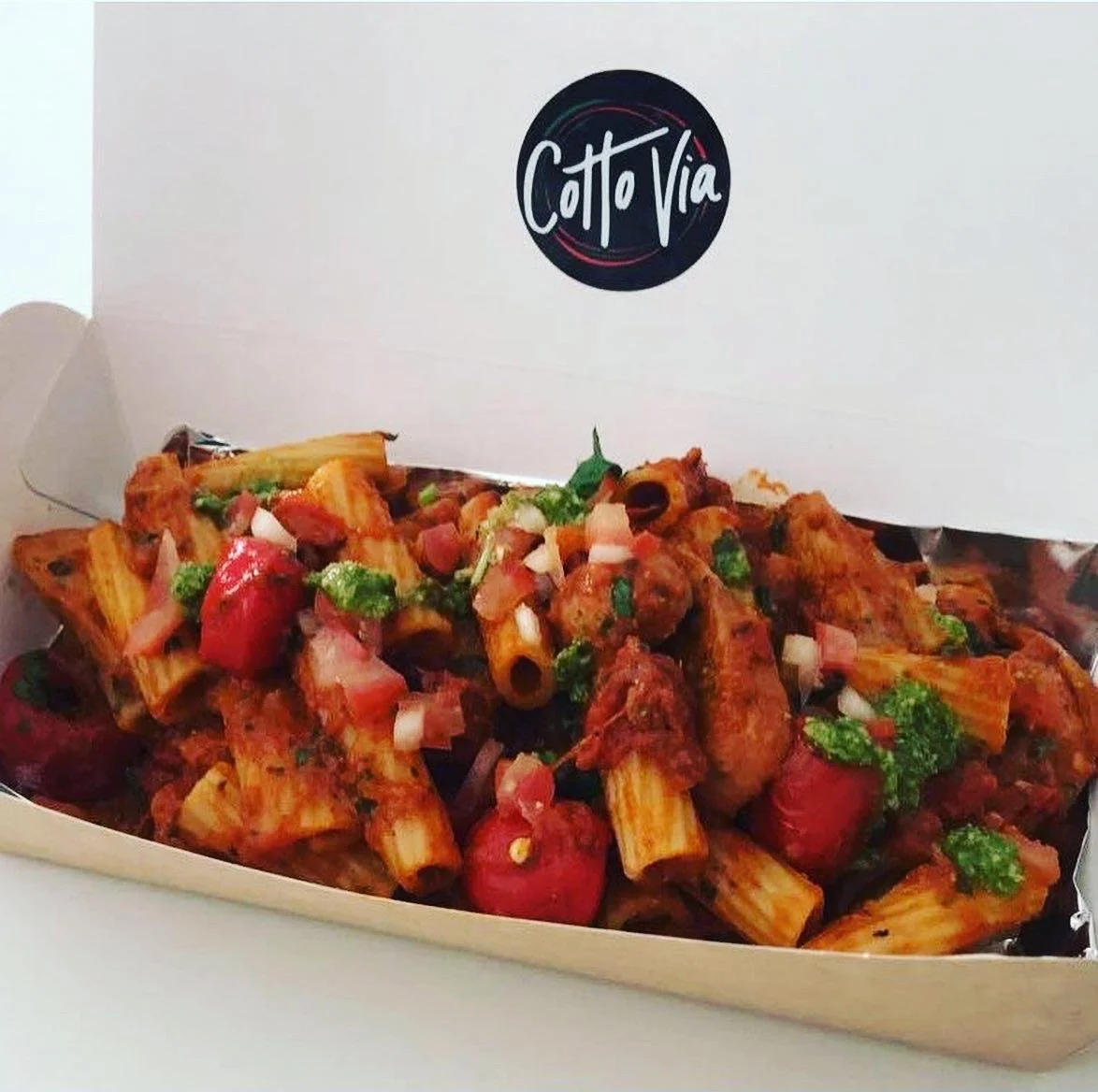 A white takeout box with a logo that says 'Cotto Via' contains pasta with tomato sauce, chopped tomatoes, onions, and green herbs.