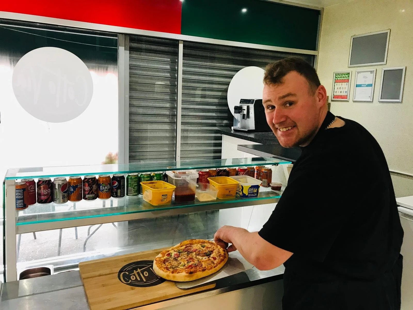 A young man with short dark hair, smiling, holding a pizza on a cutting board in a cafe or fast-food restaurant. Behind him is a glass display with canned sodas, a coffee machine, and various containers, with a closed metal shutter and a window in the background.