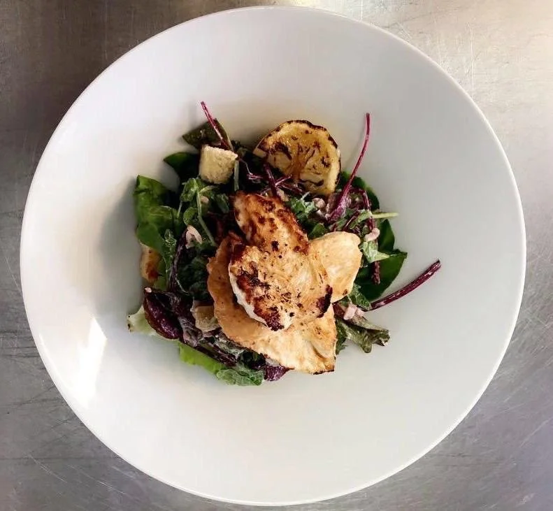 Mixed green salad with grilled chicken slices and grilled zucchini in a white bowl.