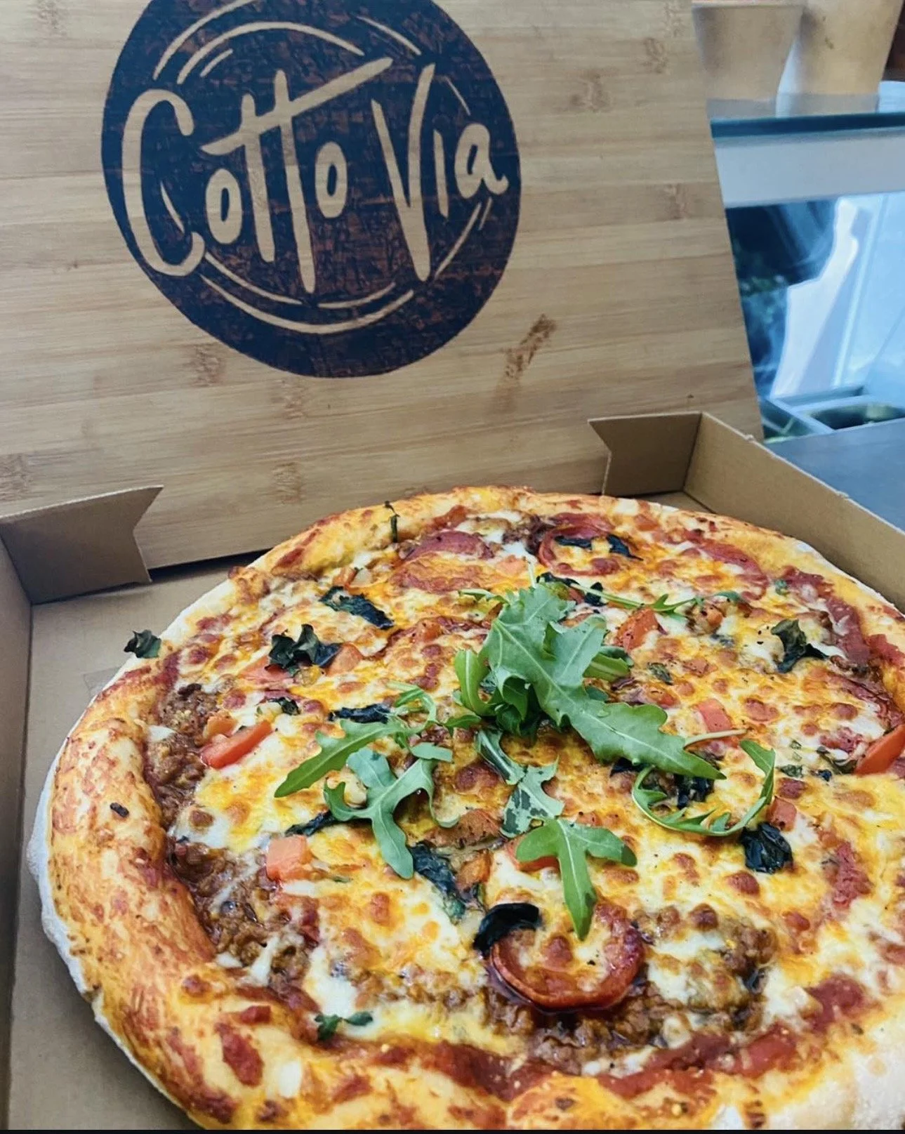 A freshly baked pizza in an open cardboard box with toppings including tomato slices, cheese, olives, and fresh arugula.