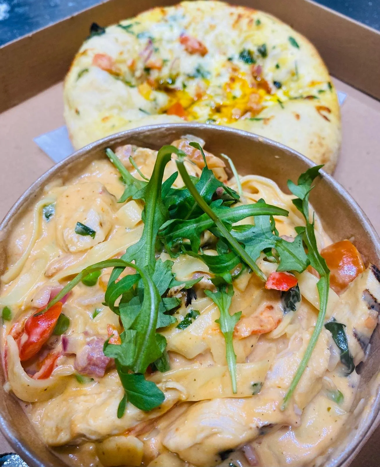 Close-up of a bowl of creamy pasta topped with fresh arugula. In the background, a pizza with toppings including cheese, vegetables, and possibly meat, in a cardboard pizza box.