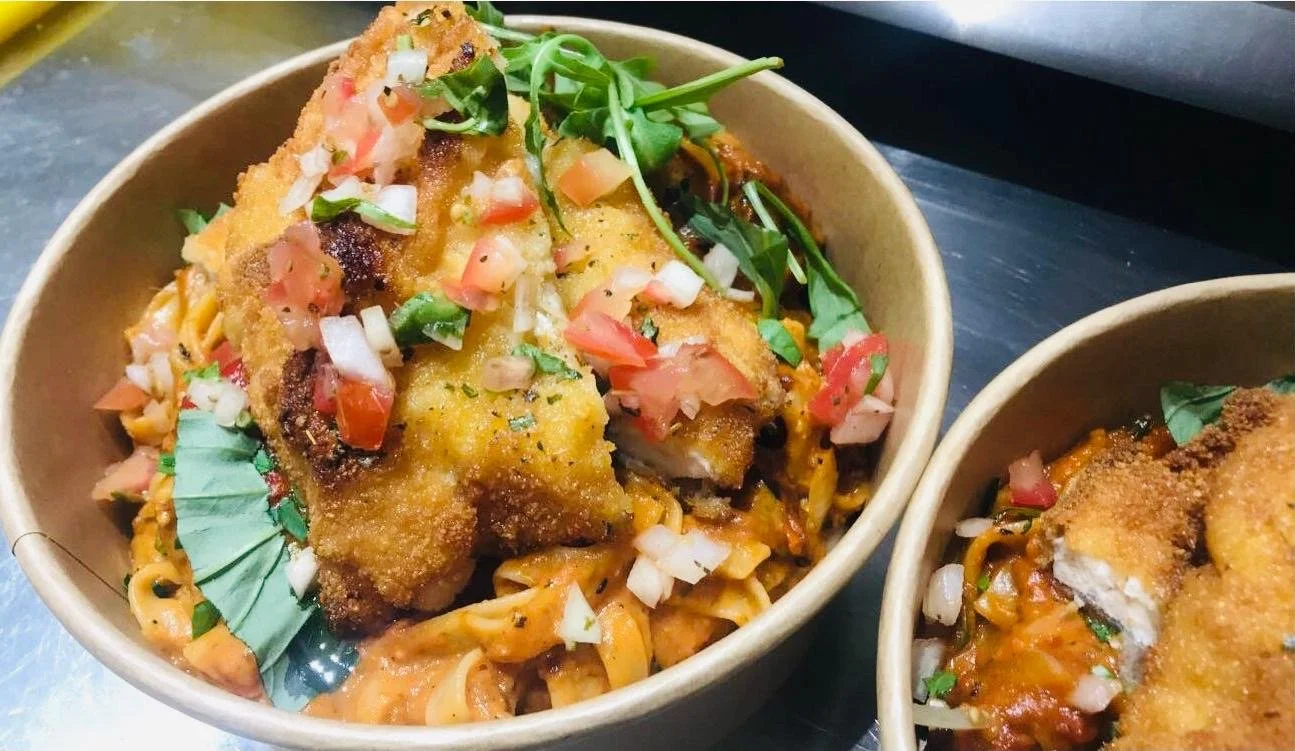 Bowl of fried fish topped with chopped tomatoes, onions, and green herbs, served over pasta with sauce.