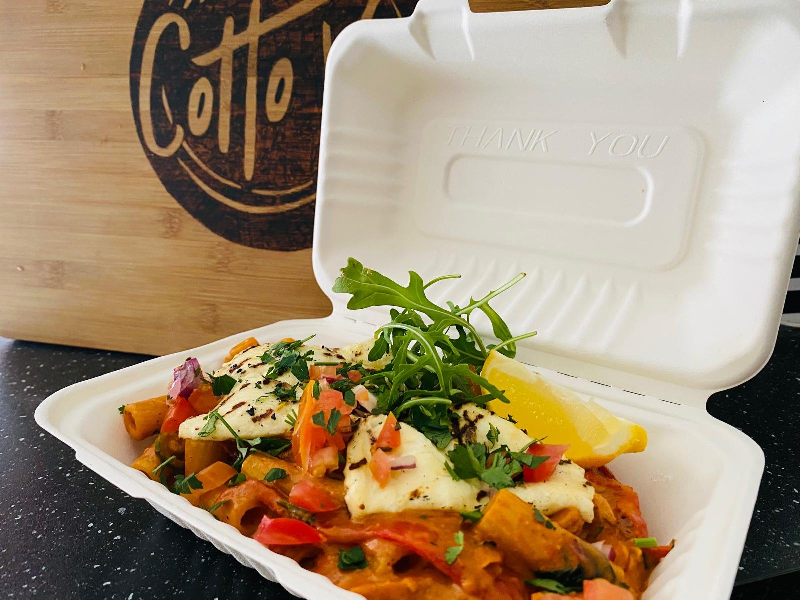 A takeout container filled with a colorful dish topped with grilled chicken, fresh herbs, lemon wedge, and fresh greens on top. In the background, there is a wooden coffee sign.