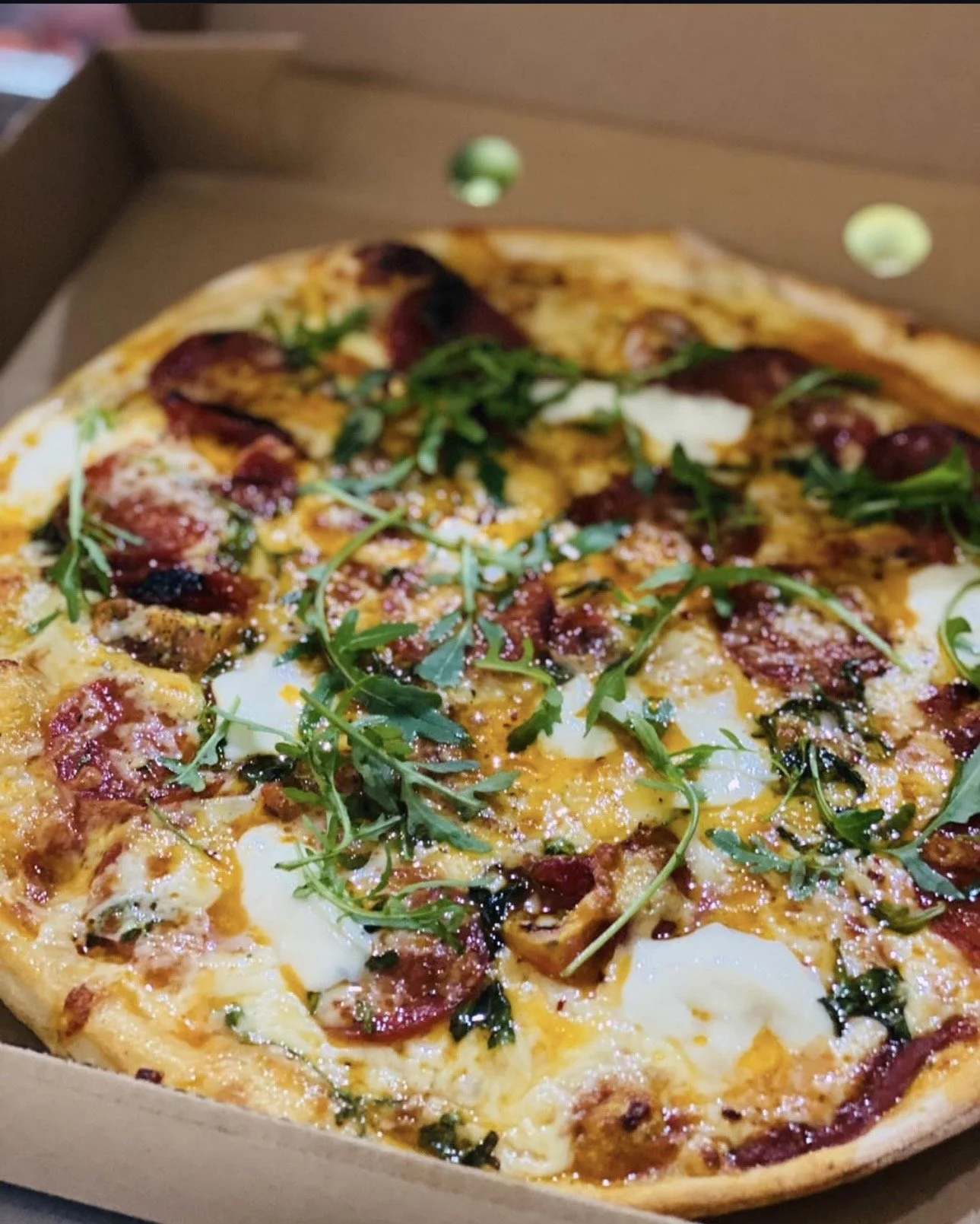 A pizza with pepperoni, mozzarella cheese, arugula, and a sprinkle of grated cheese in a cardboard box.