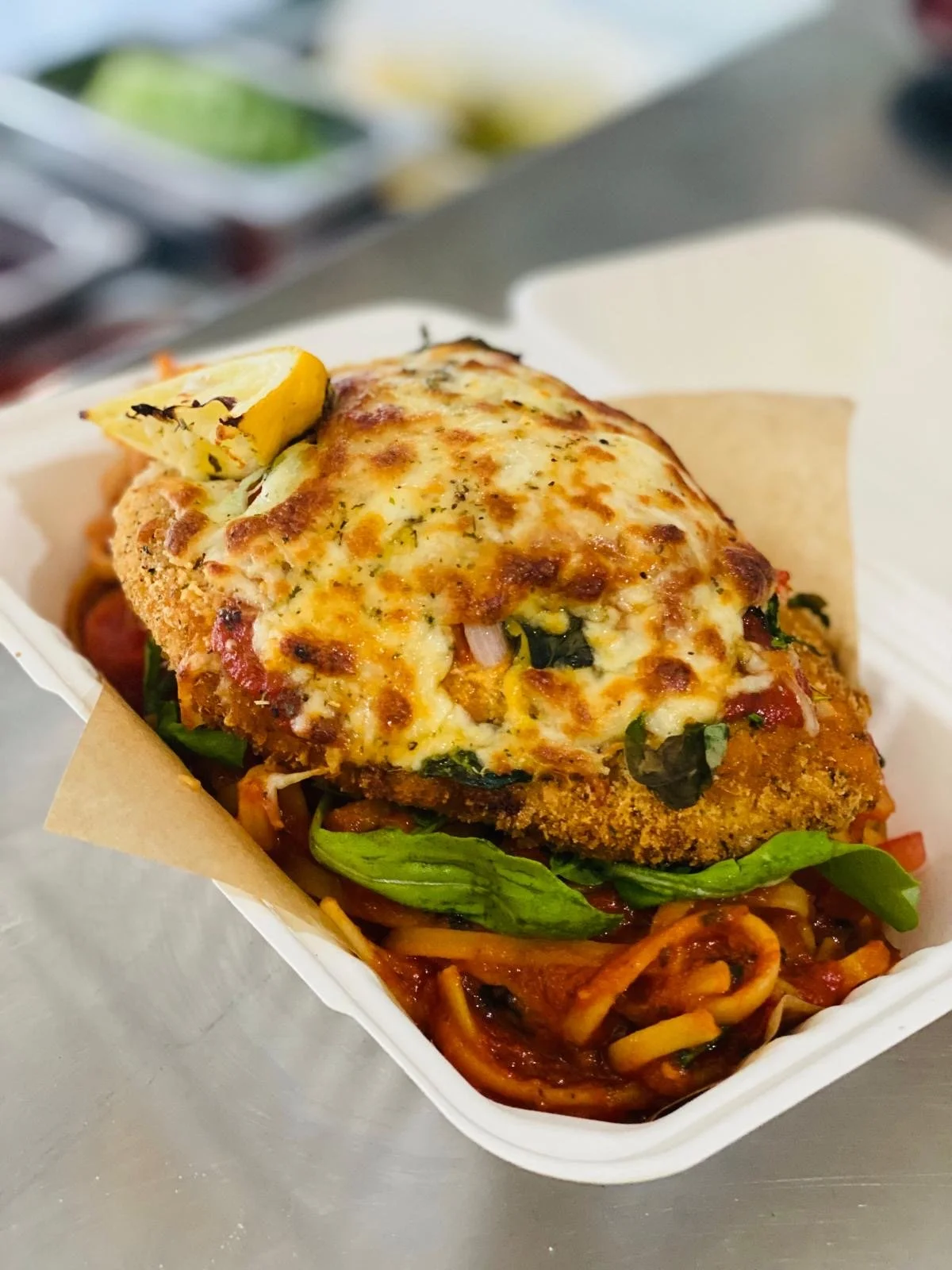 A breaded chicken breast topped with melted cheese and a lemon wedge served on a bed of spaghetti with tomato sauce, vegetables, and fresh greens.