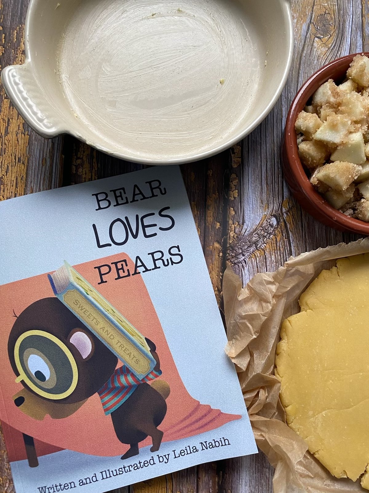 A book titled "Bear Loves Pears" by Leila Nabih, a partially visible empty baking bowl, on a wooden surface.