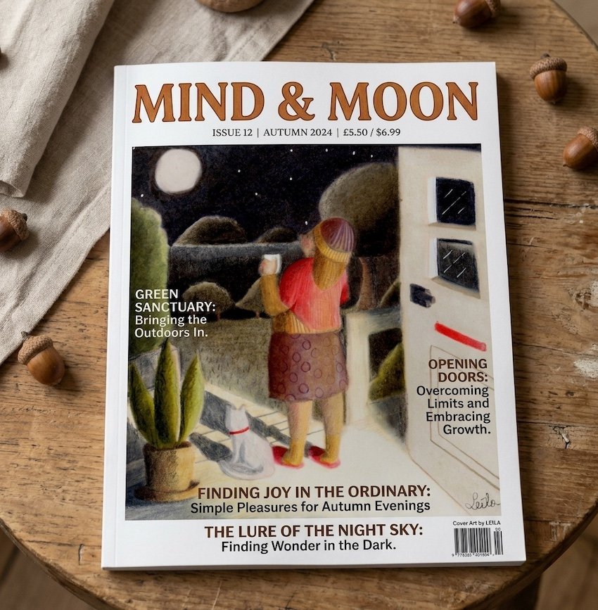 A photograph of the 'Mind & Moon' magazine Autumn 2024 cover (Issue 12), resting on a rustic wooden table with acorns. The cover features a colored pencil illustration of a woman and a cat at an open doorway, looking out at a full moon under a starry
