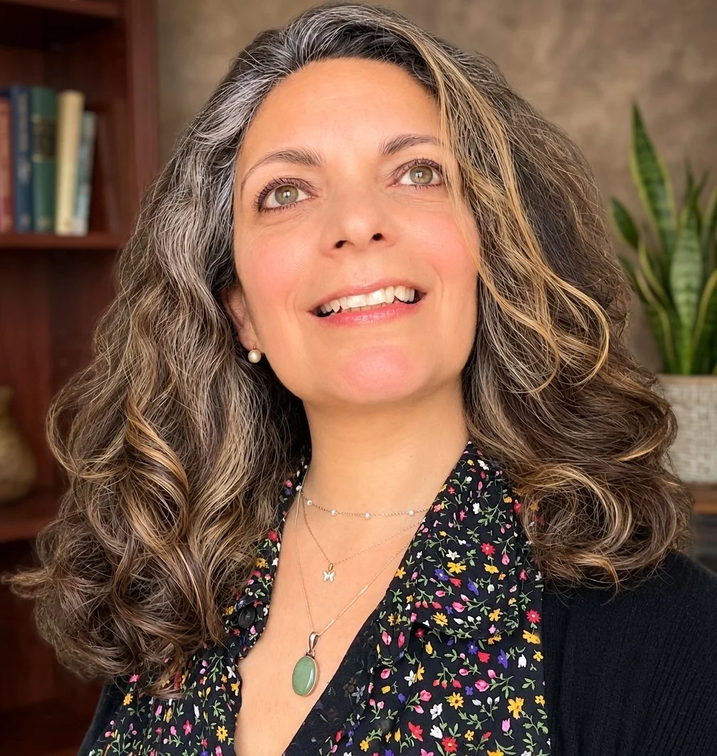 Smiling woman with long wavy gray-streaked hair wearing a floral blouse and layered necklaces indoors